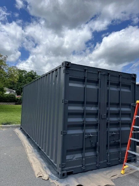 A large, dark gray shipping container placed on a paved surface outdoors under a partly cloudy sky, with a ladder leaning against it and a strip of fabric or tarp on the ground nearby.
