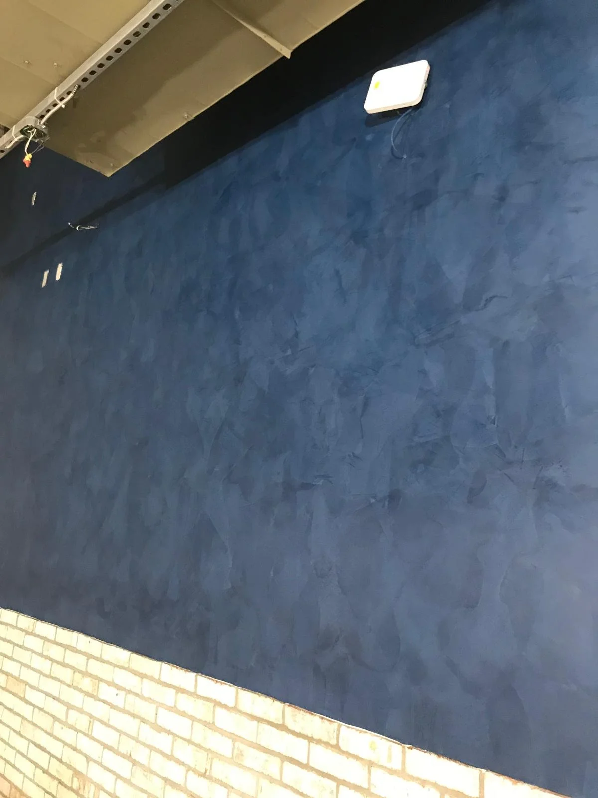 A partially finished interior wall with a blue textured surface and a section of light-colored brick at the bottom. There are wires and a white electronic device mounted on the wall.