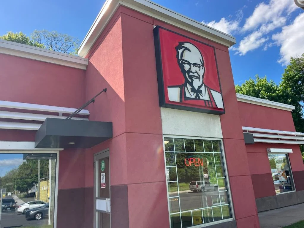 Exterior of a KFC fast-food restaurant with a large Colonel Sanders logo sign on a red building. The window displays a neon 'OPEN' sign, and the sky is blue with some clouds.