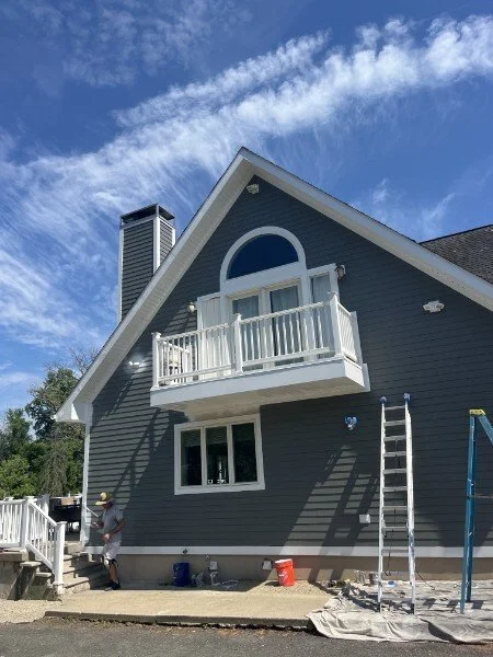 A two-story blue house with a small balcony on the second floor, under a partly cloudy sky. There is a ladder leaning against the house and construction materials nearby, indicating ongoing work.