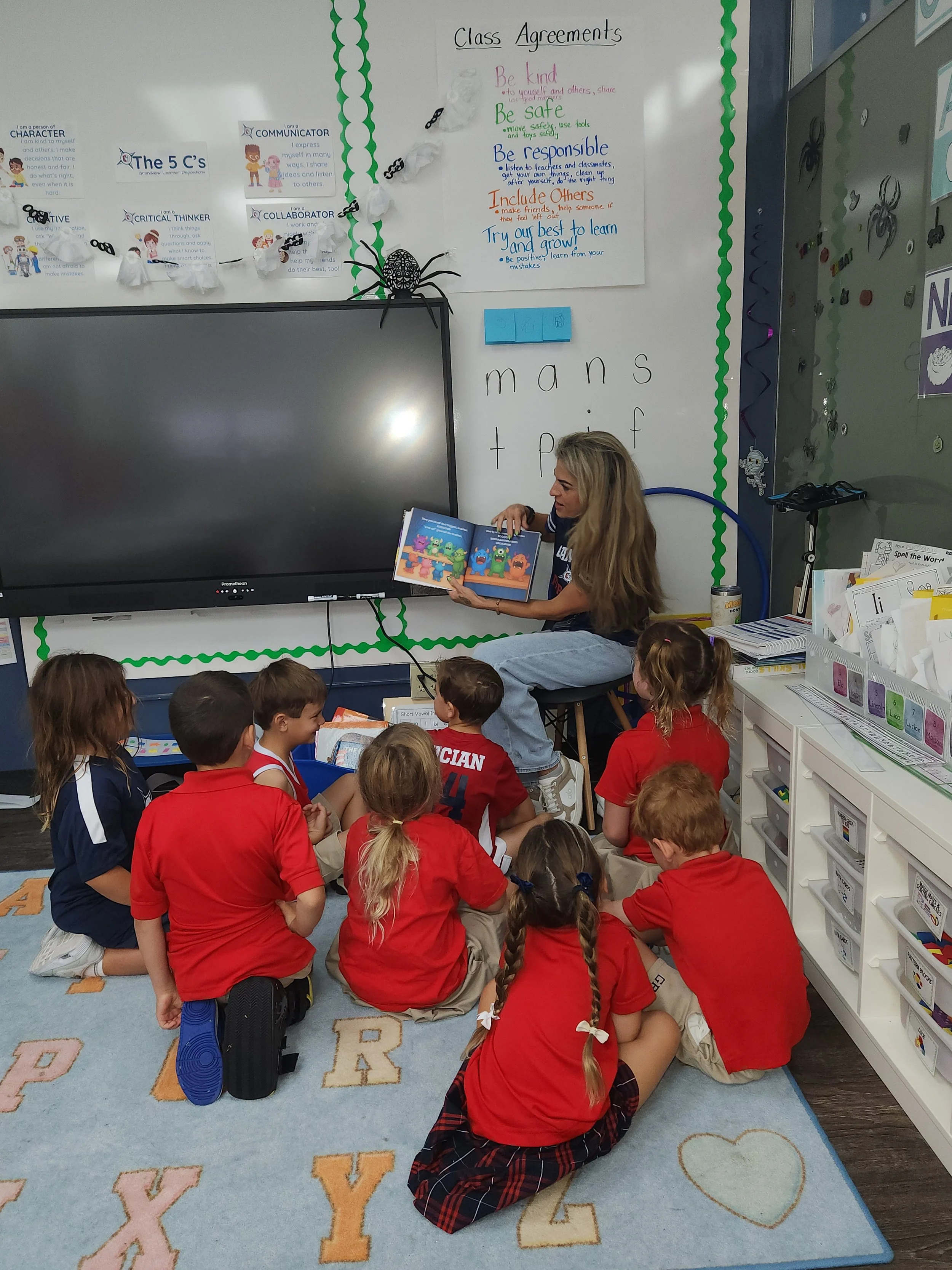 A teacher reading a colorful children’s book to a group of students seated on the carpet in a classroom. The students are wearing red and blue uniforms, and the classroom decor includes educational posters and books.
