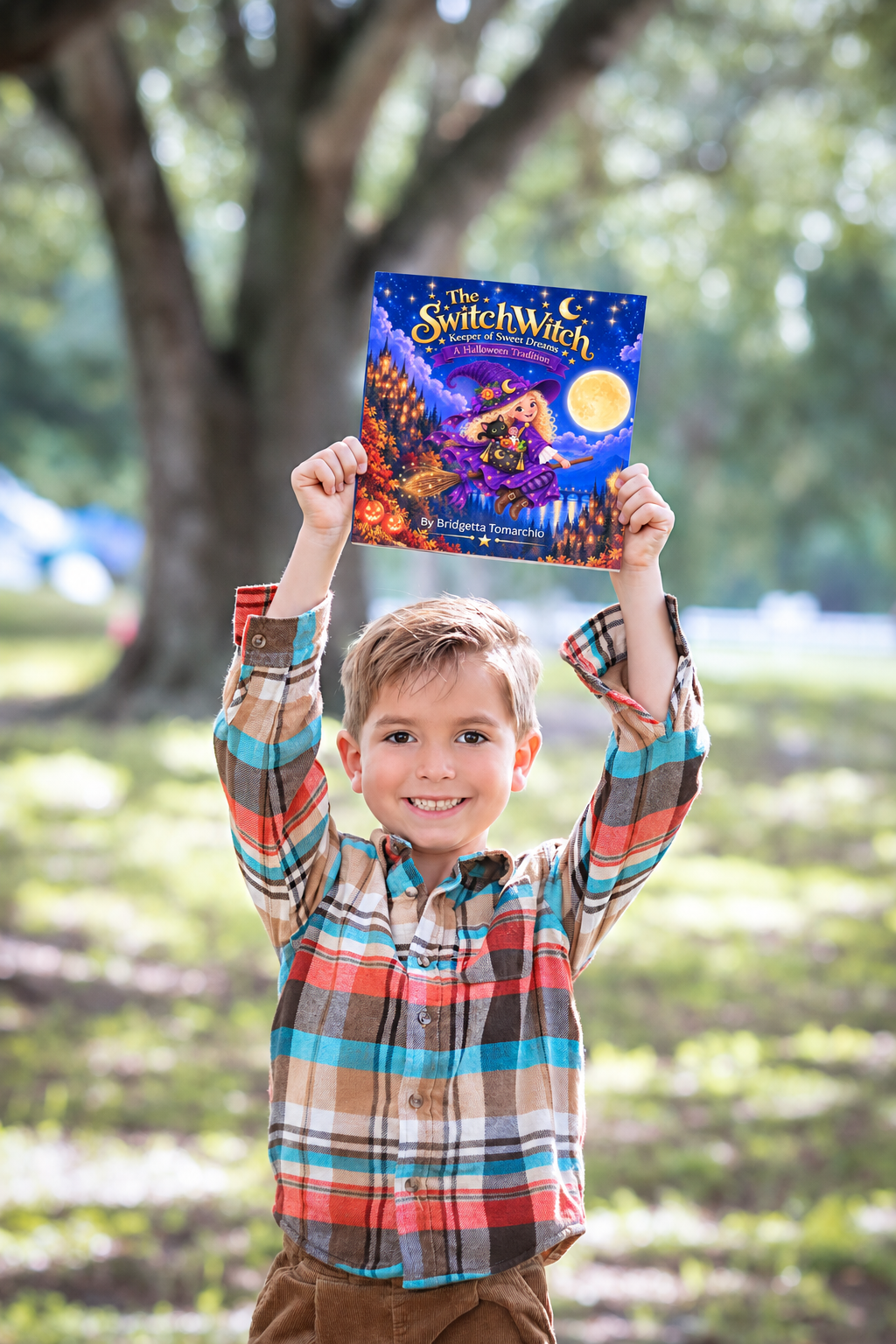 A young boy with a big smile, wearing a plaid shirt, holds up a colorful book titled "The Switch Witch" outdoors in front of a large tree.