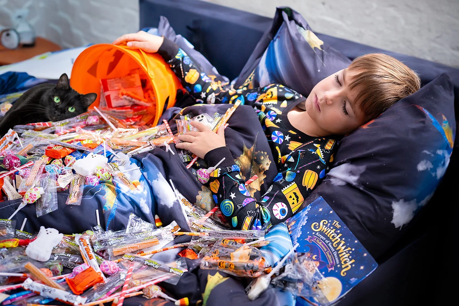 A young boy sleeping in bed surrounded by scattered candies and a black cat lying nearby with bright green eyes.