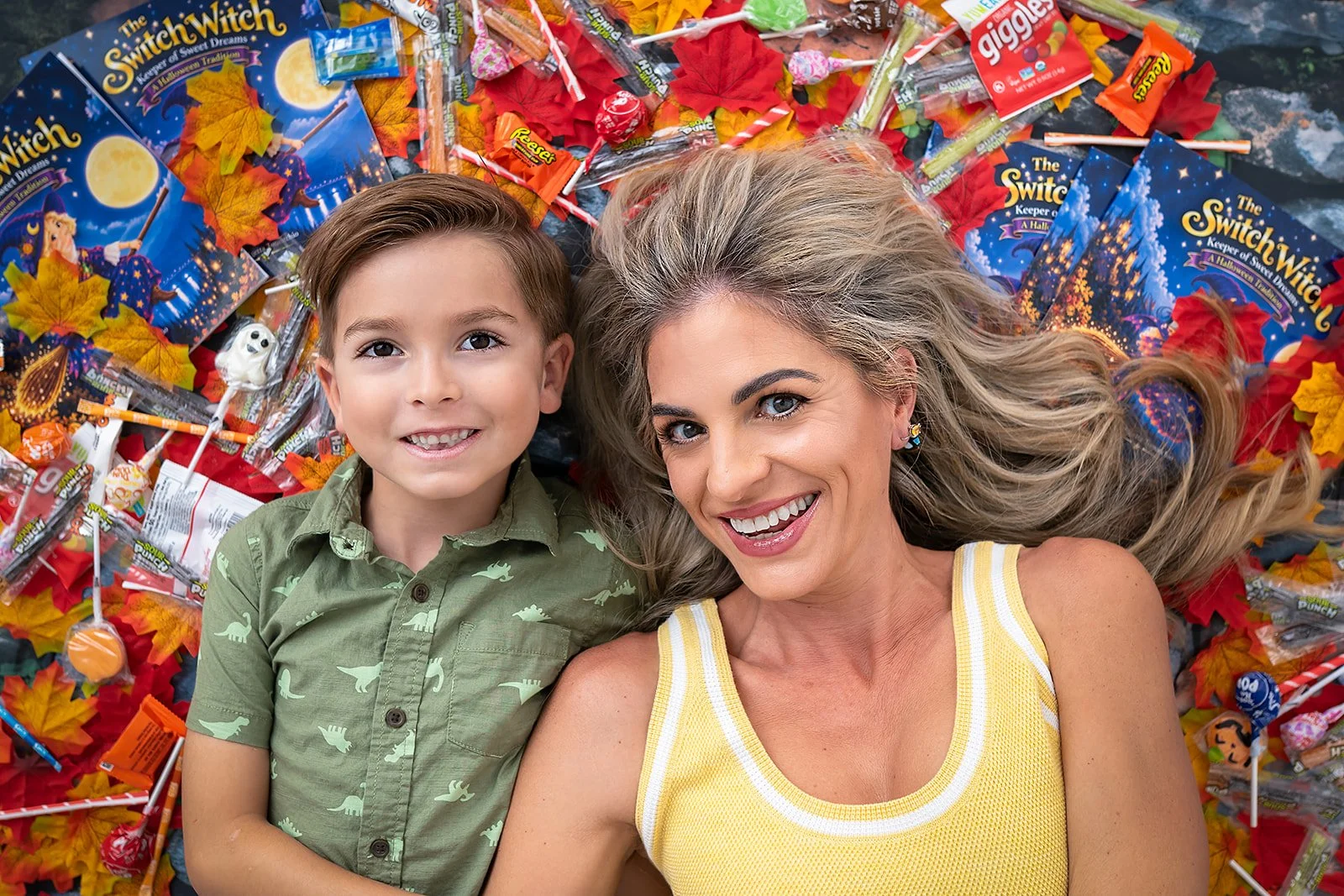 A smiling woman with blonde hair lying next to a smiling boy with brown hair, both lying on a bed of Halloween-themed candies, leaves, and Halloween decorations.