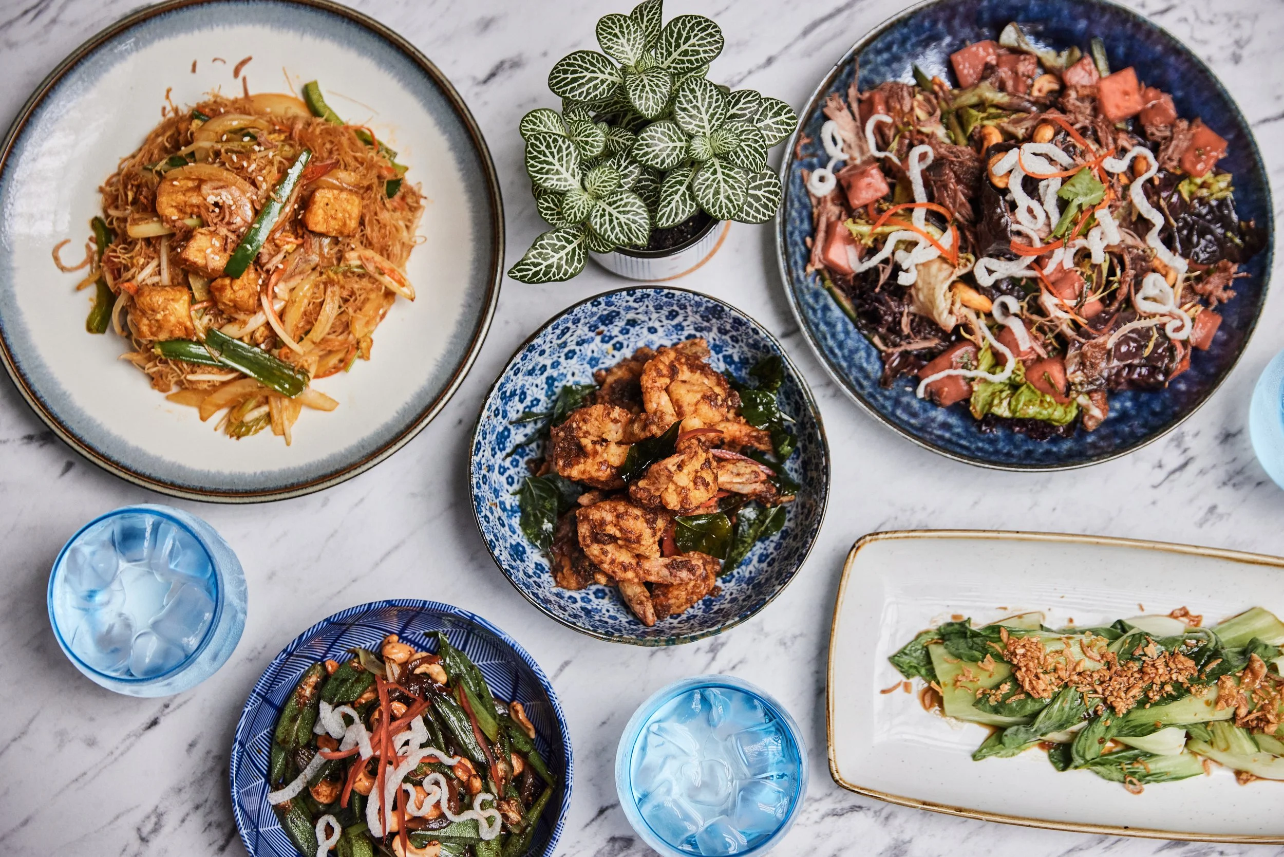 Overhead view of an Asian dinner spread at t HUŎ with stir-fried noodles, mixed vegetables, kam heong prawns, and iced drinks on a marble table