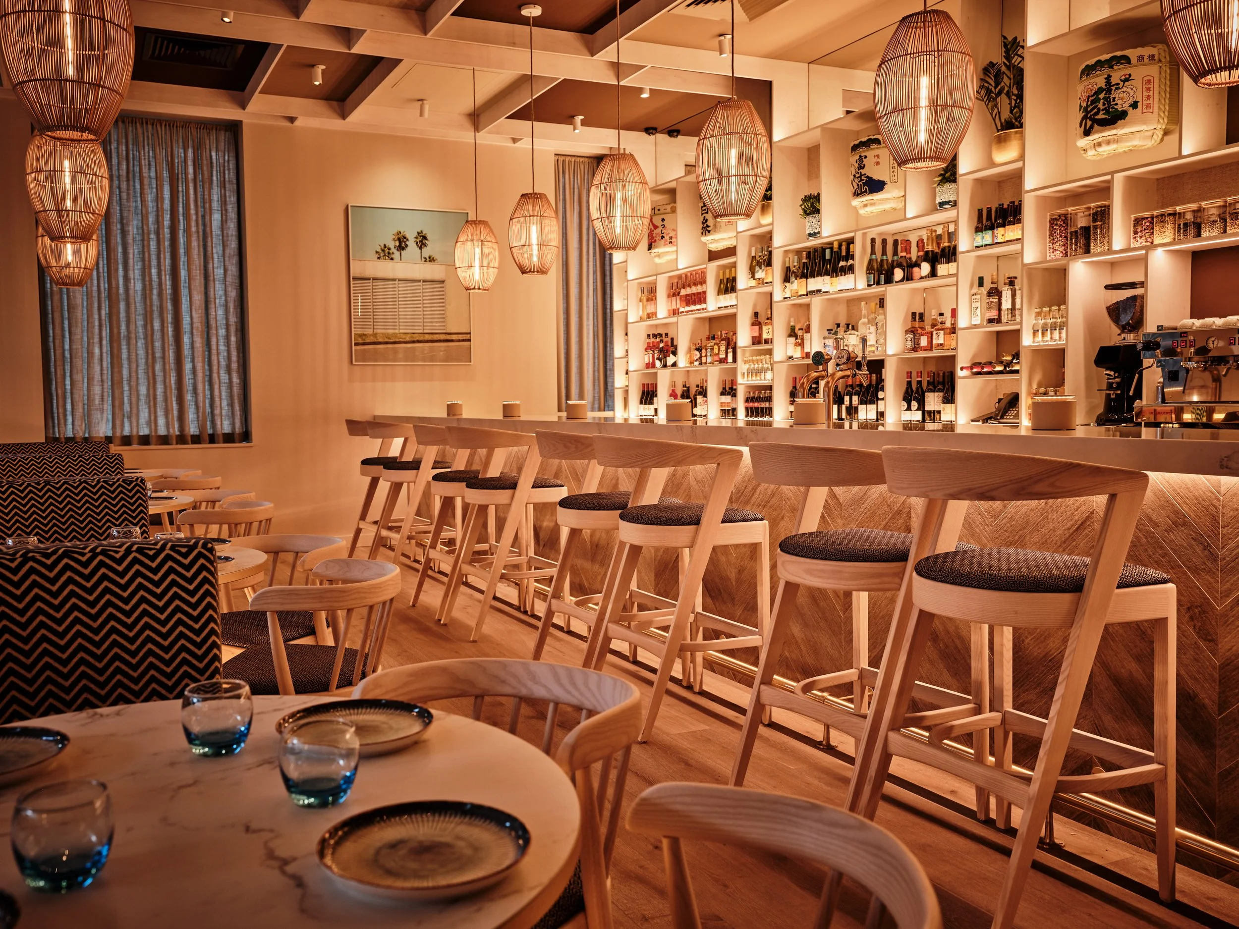 Warmly lit Asian restaurant HUŎ Belsize Village bar with woven pendant lights, wooden seating, liquor shelves, and set dining tables.