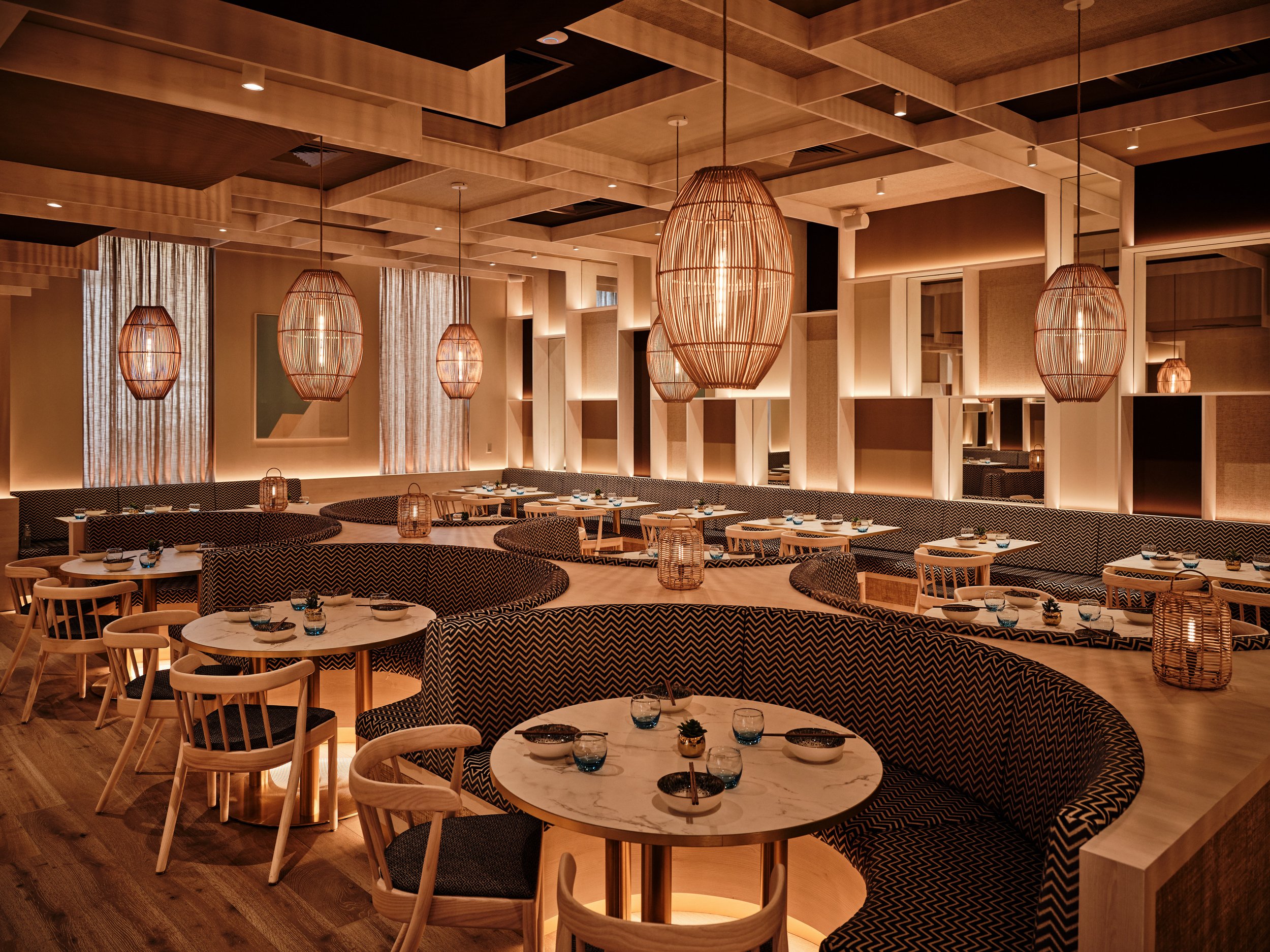Warmly lit modern restaurant interior of HUŎ Belsize Village with patterned banquette seating, round dining tables, and woven pendant lights