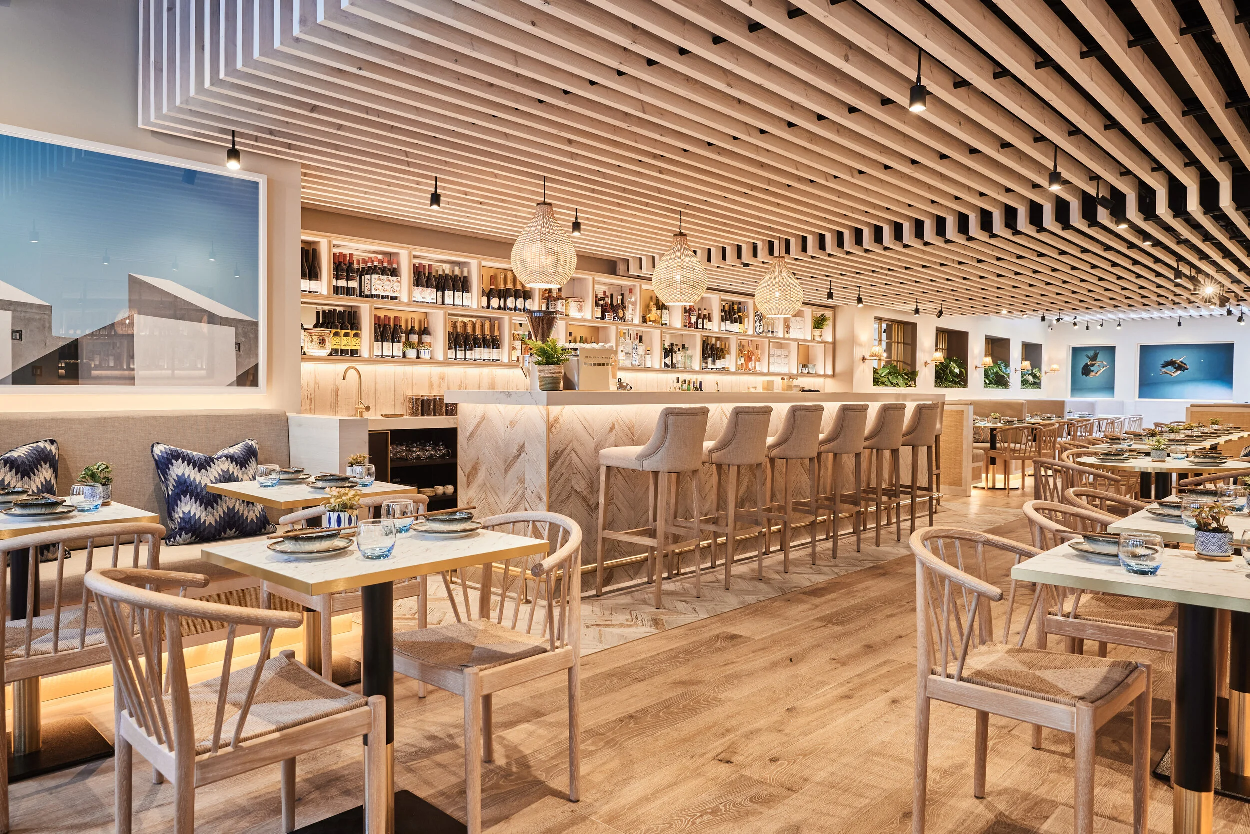 Bright modern restaurant interior of HUŎ Chelsea with a stocked wine bar, pendant lights, wood slat ceiling, seated dining tables, and bar stools