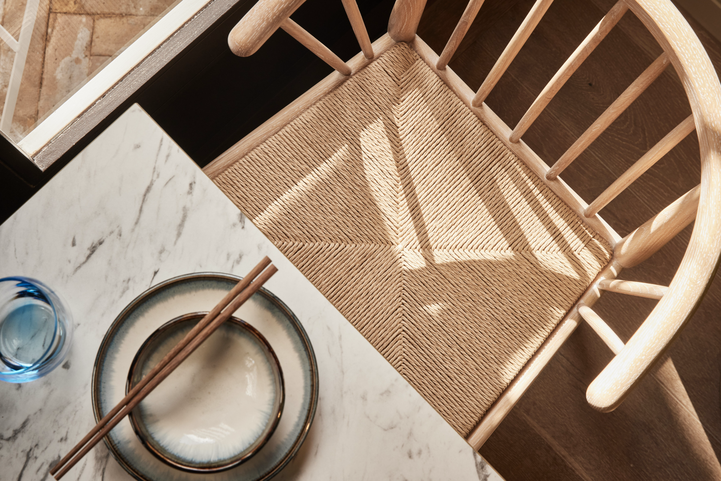 Top-down view of a woven wooden dining chair at HUŎ beside a marble table set with a plate, chopsticks, and blue drinking glass