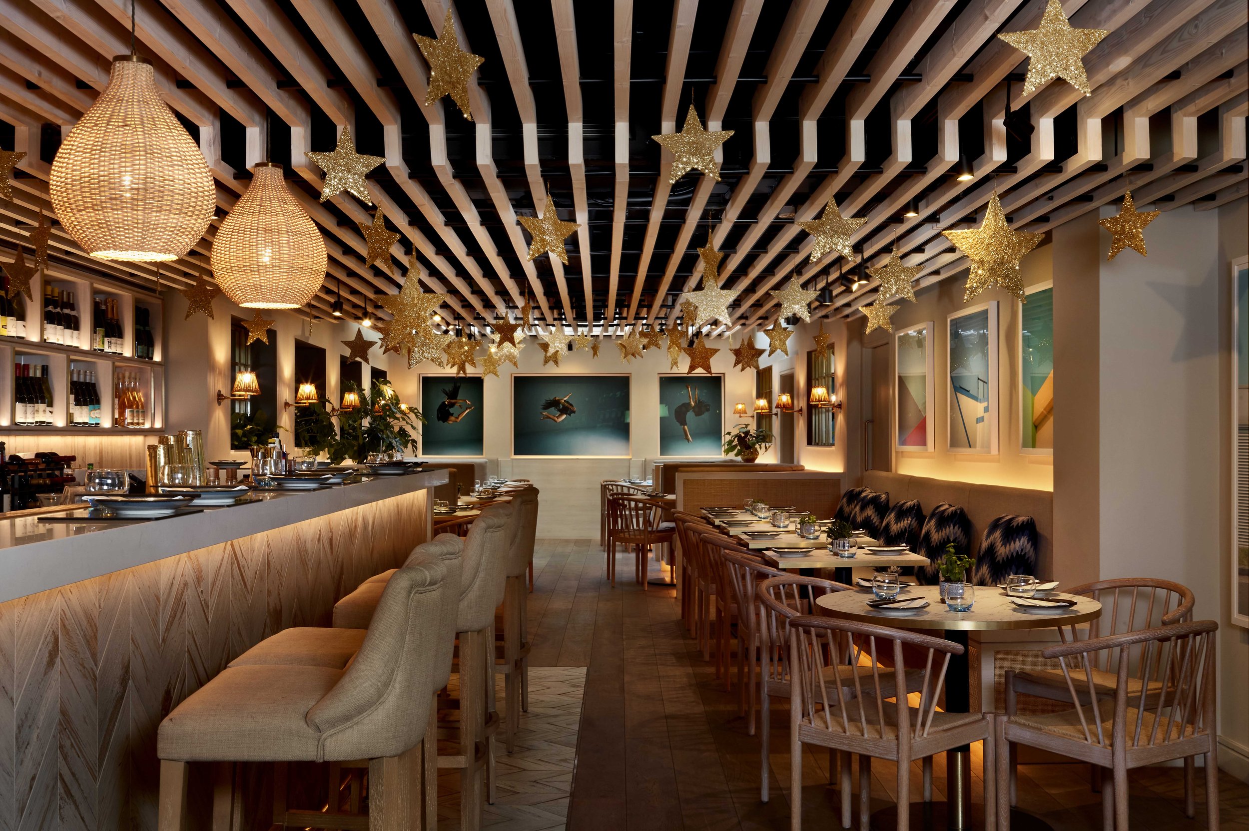 Warmly lit restaurant interior of HUŎ Chelsea with a bar, dining tables, woven pendant lights, and gold star decorations hanging from a wood-slat ceiling