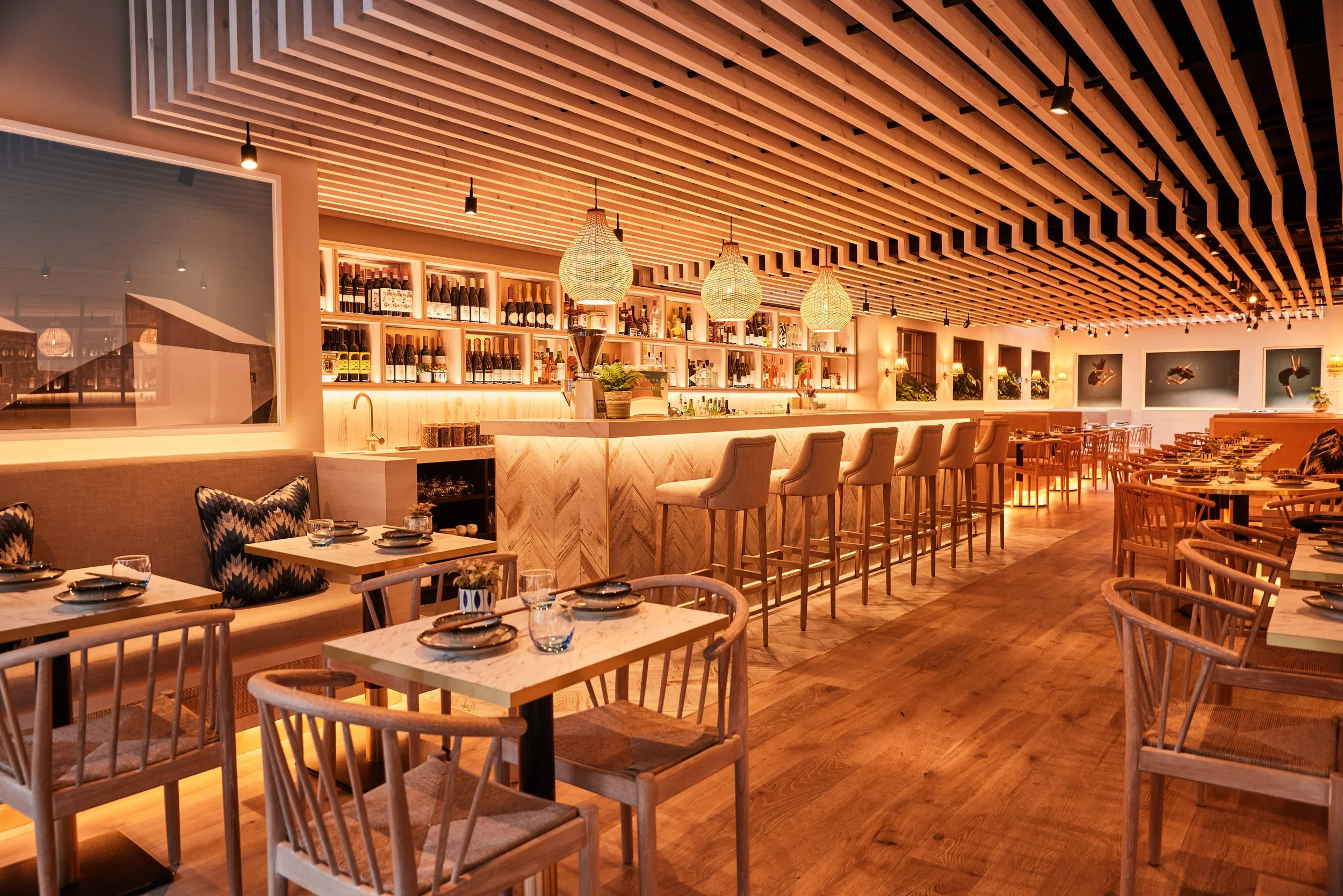 Warmly lit interior of HUŎ Chelsea upscale restaurant interior with a stocked bar, wicker pendant lights, wood slat ceiling, and neatly set dining tables.
