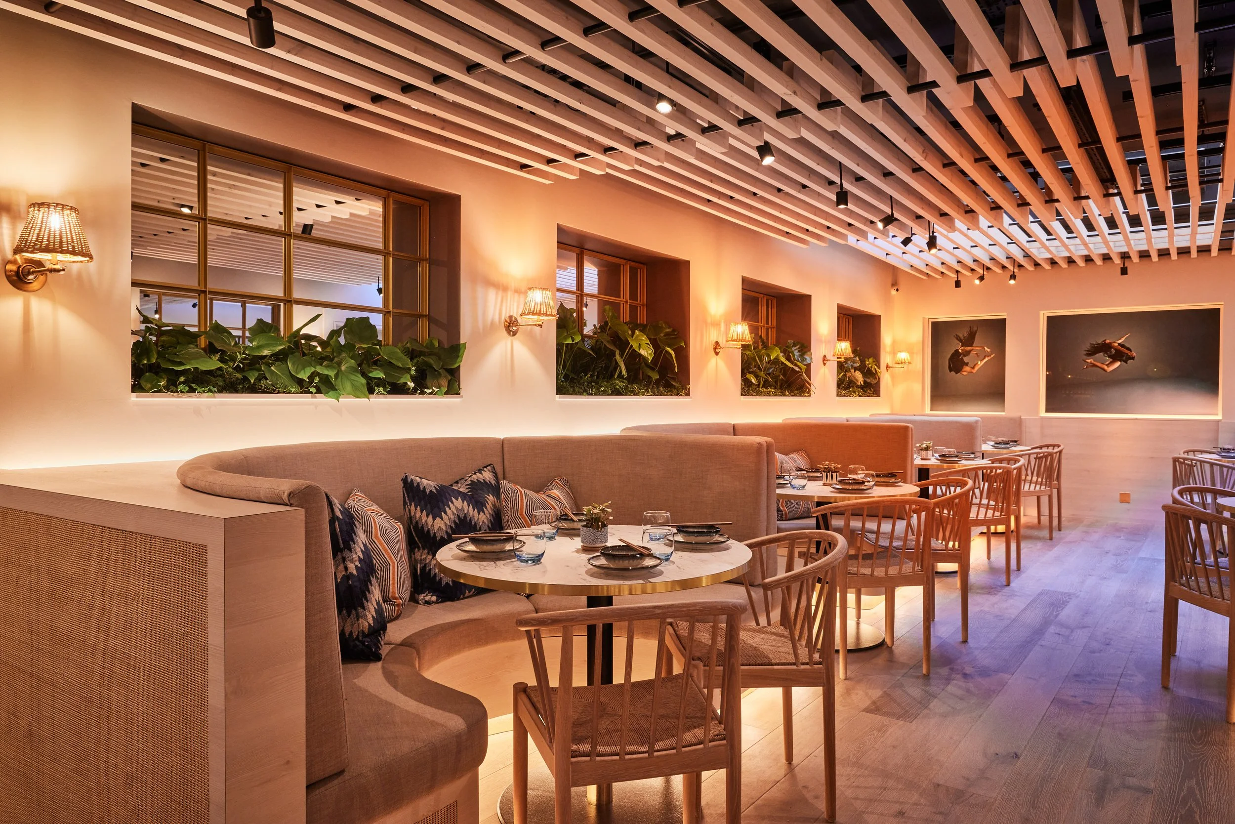 Warmly lit modern restaurant interior of HUŎ Chelsea with upholstered booths, round dining tables, wooden chairs, plants, wall sconces, and a slatted wood ceiling