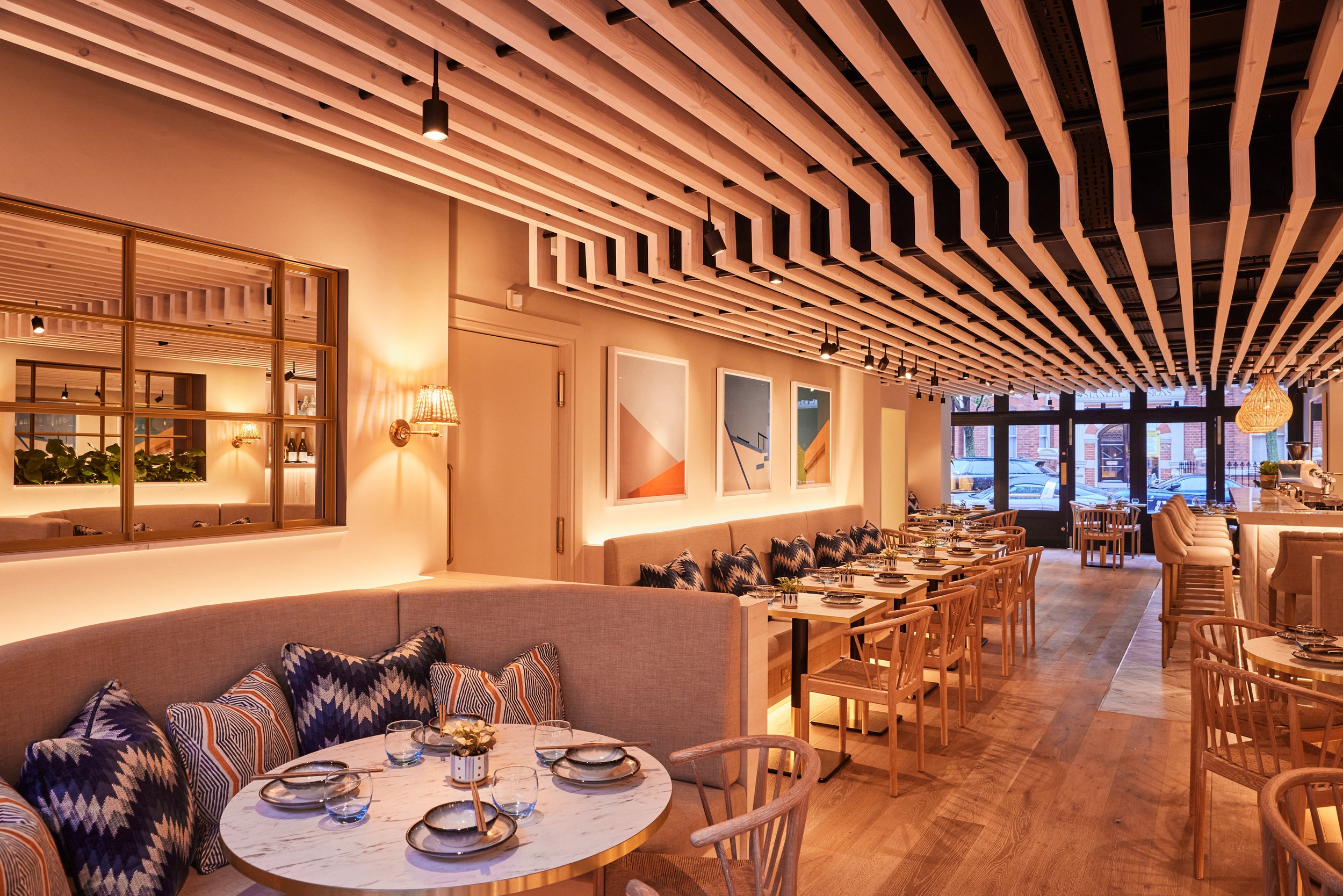 Warmly lit modern restaurant interior of HUŎ Chelsea with wood slat ceiling, dining tables, upholstered booths, patterned pillows, and pendant lighting