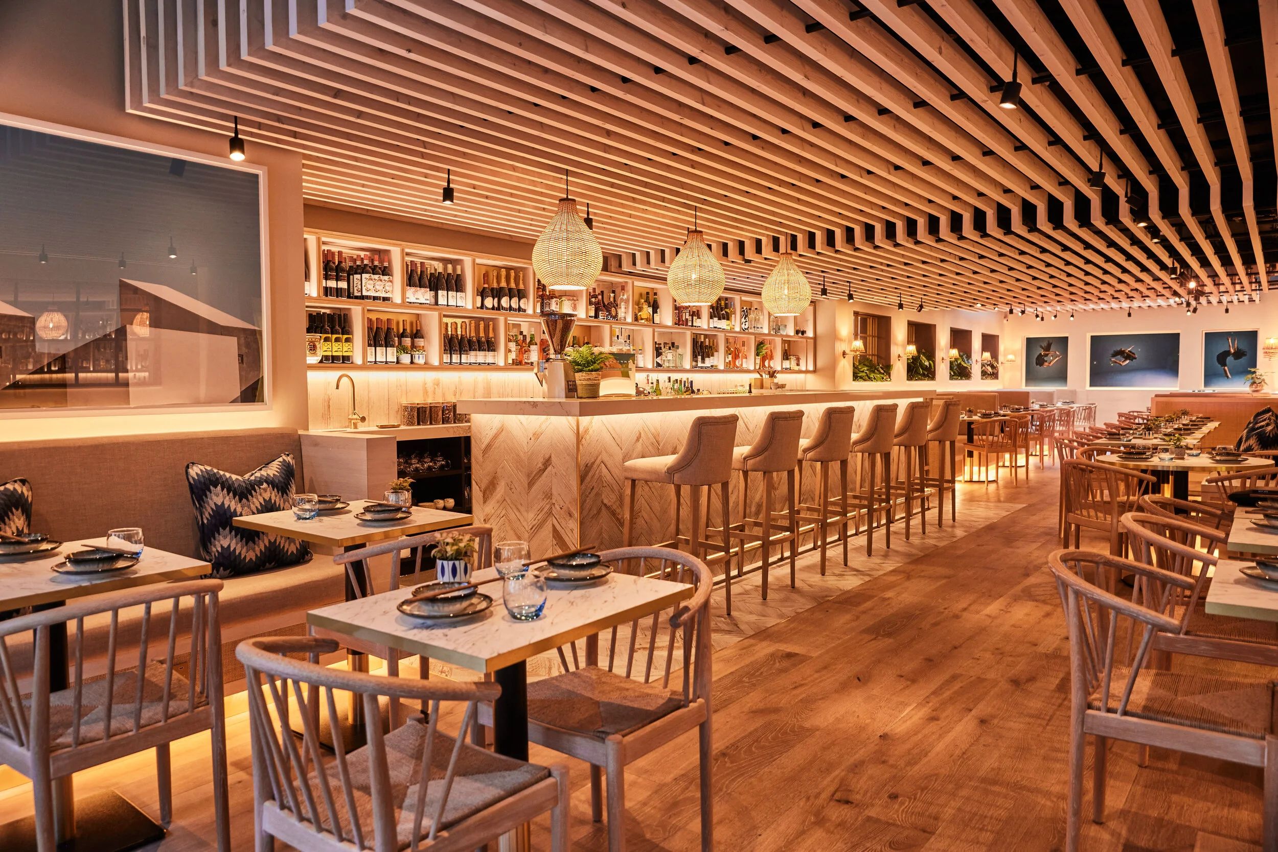 Warmly lit modern restaurant interior of HUŎ Chelsea with a stocked bar, pendant lights, wood slat ceiling, and neatly set dining tables.