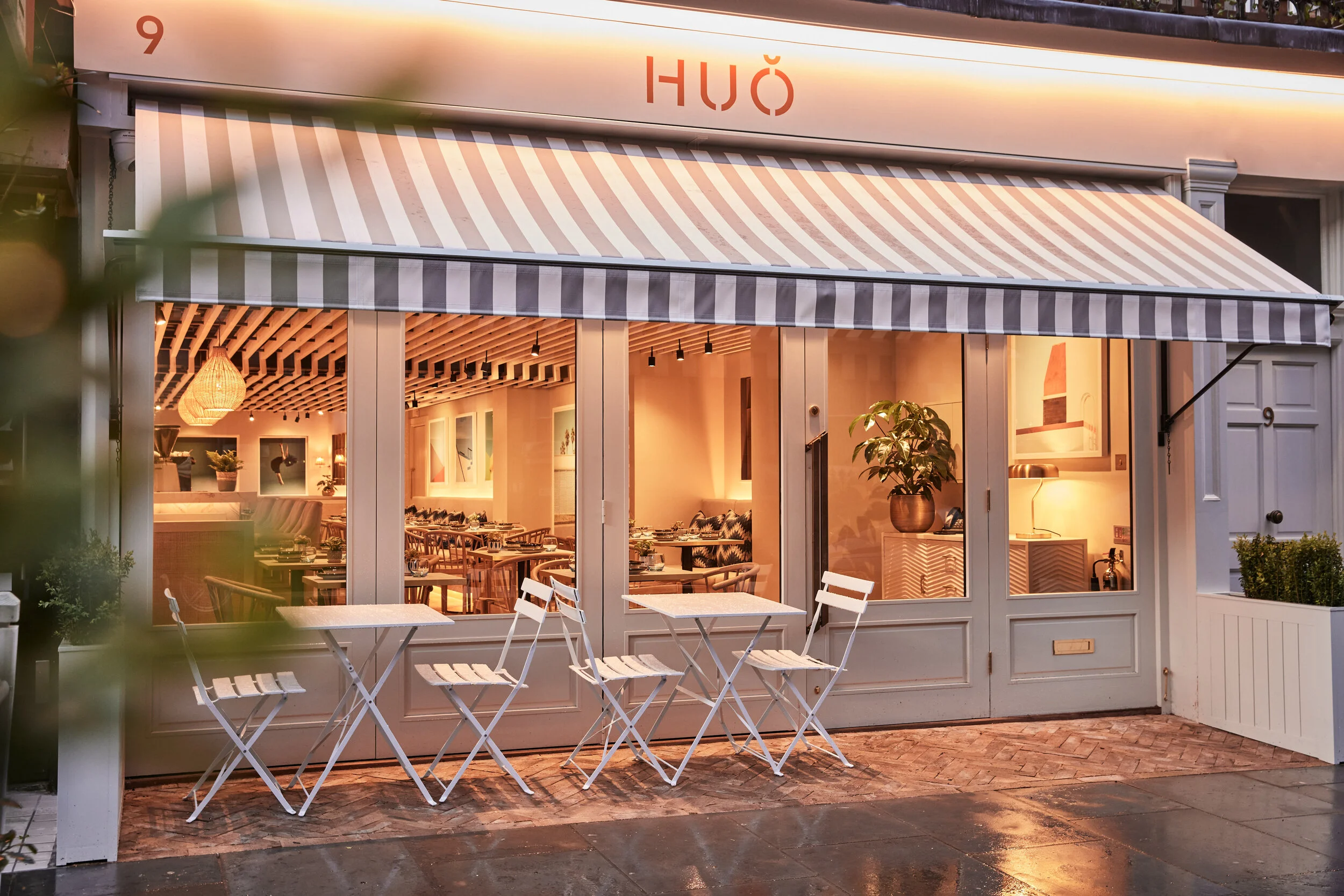 Exterior of HUŎ restaurant with striped awning, illuminated dining room, and outdoor café tables and chairs.