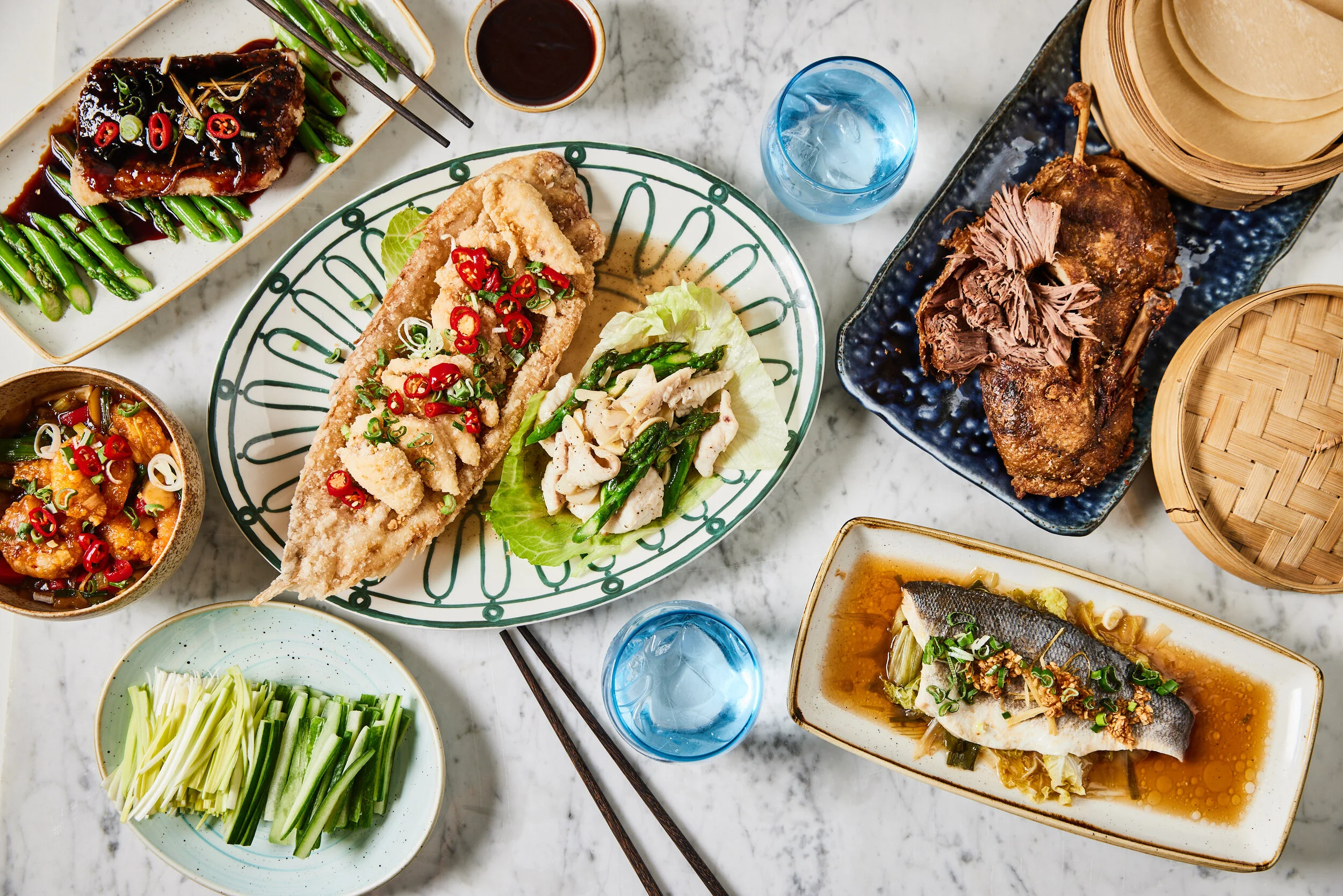 Overhead spread at HUŎ Asian dishes, including crispy fish, glazed meat, roasted duck, steamed fish, stir-fried vegetables, dipping sauce, chopsticks, and bamboo steamers.