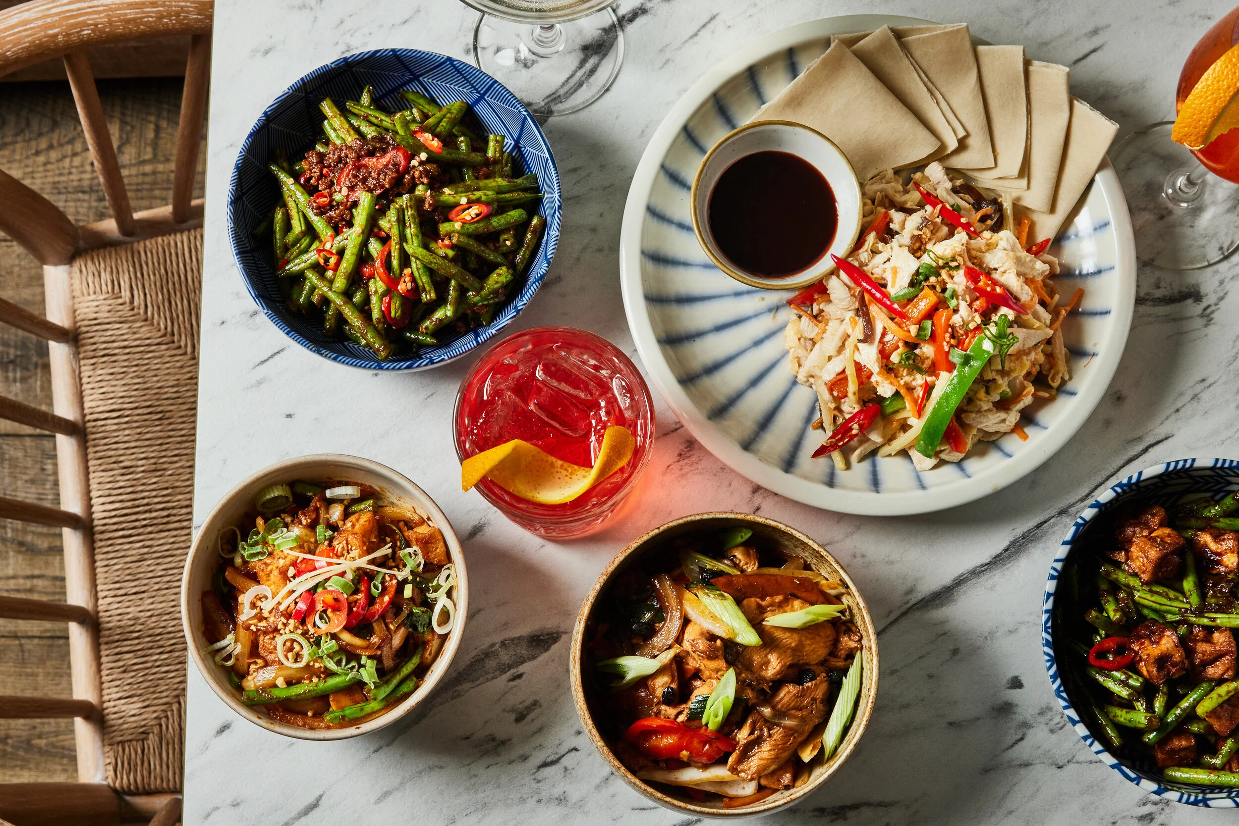 Overhead view of a spread of food at HUŎ with spicy green beans, stir-fried chicken and vegetables, spring rolls, dipping sauce, and citrus cocktails.