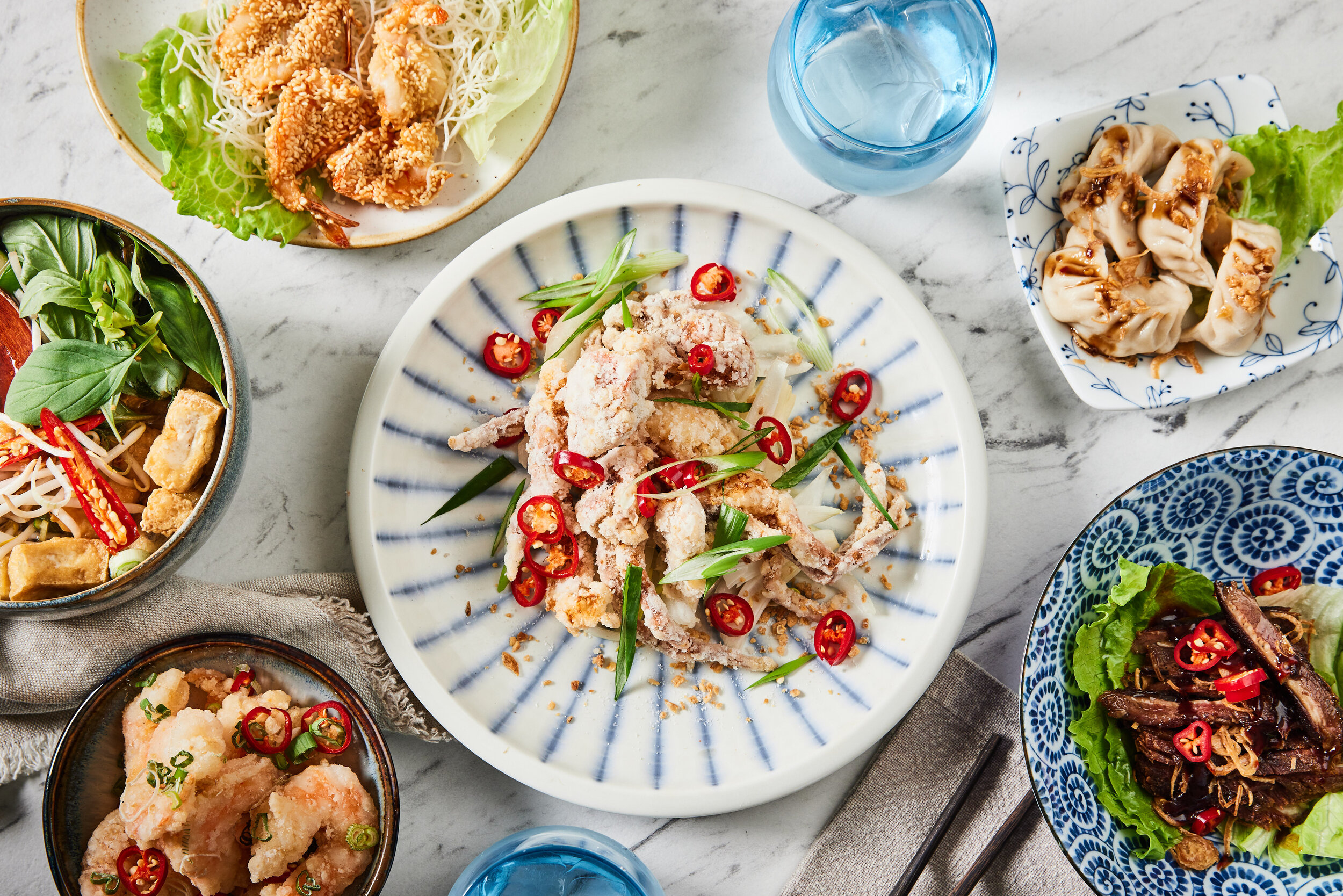 Overhead spread of Asian dishes at HUŎ, including fried seafood, sesame shrimp, dumplings, noodles, and sliced steak with chili peppers