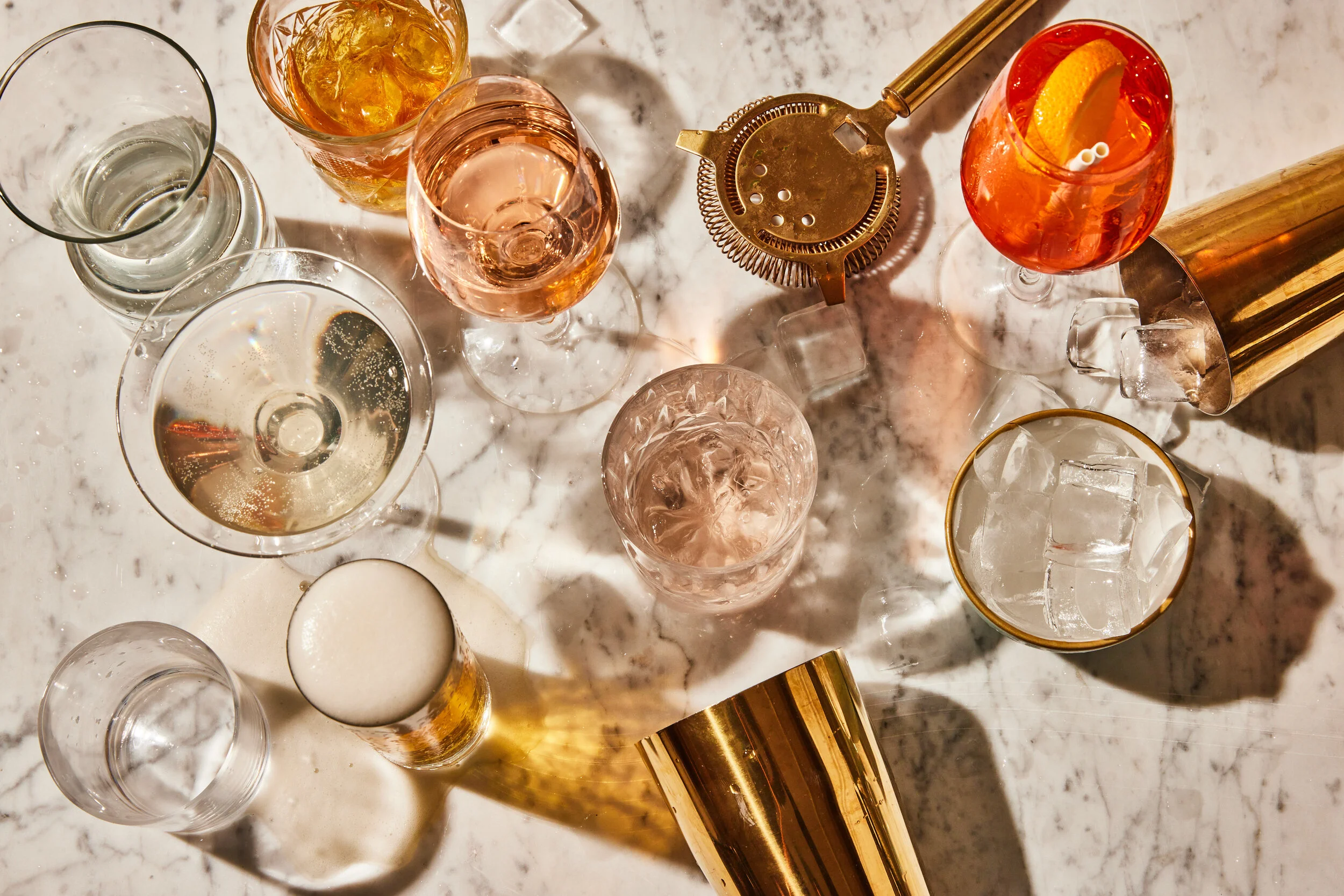 Overhead view of assorted cocktails, liquor glasses, ice, orange garnishes, and gold bar tools on a marble countertop at HUŎ