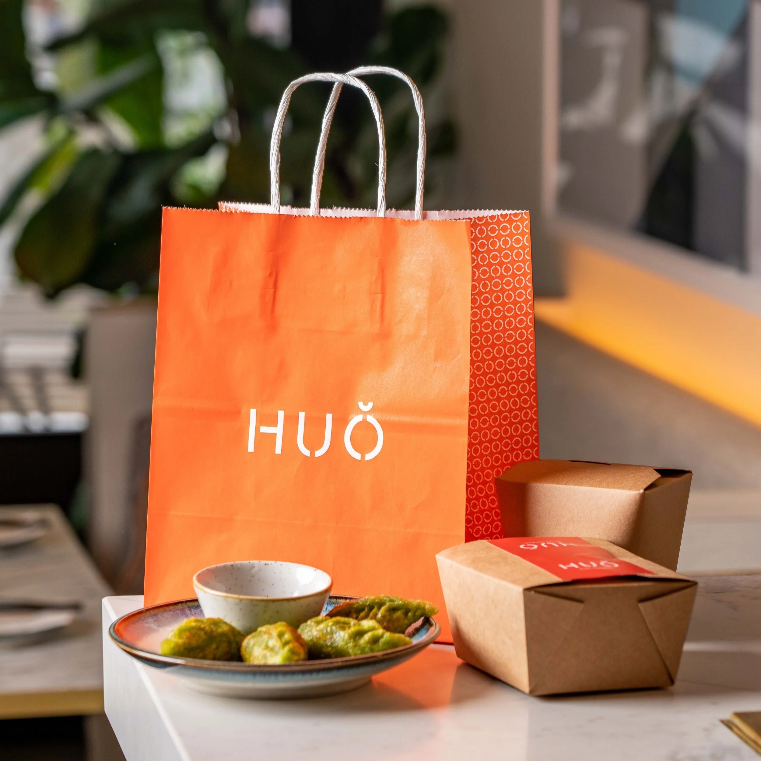 Orange HUŎ shopping bag, packaged food boxes, and a plate of green gyoza on a table
