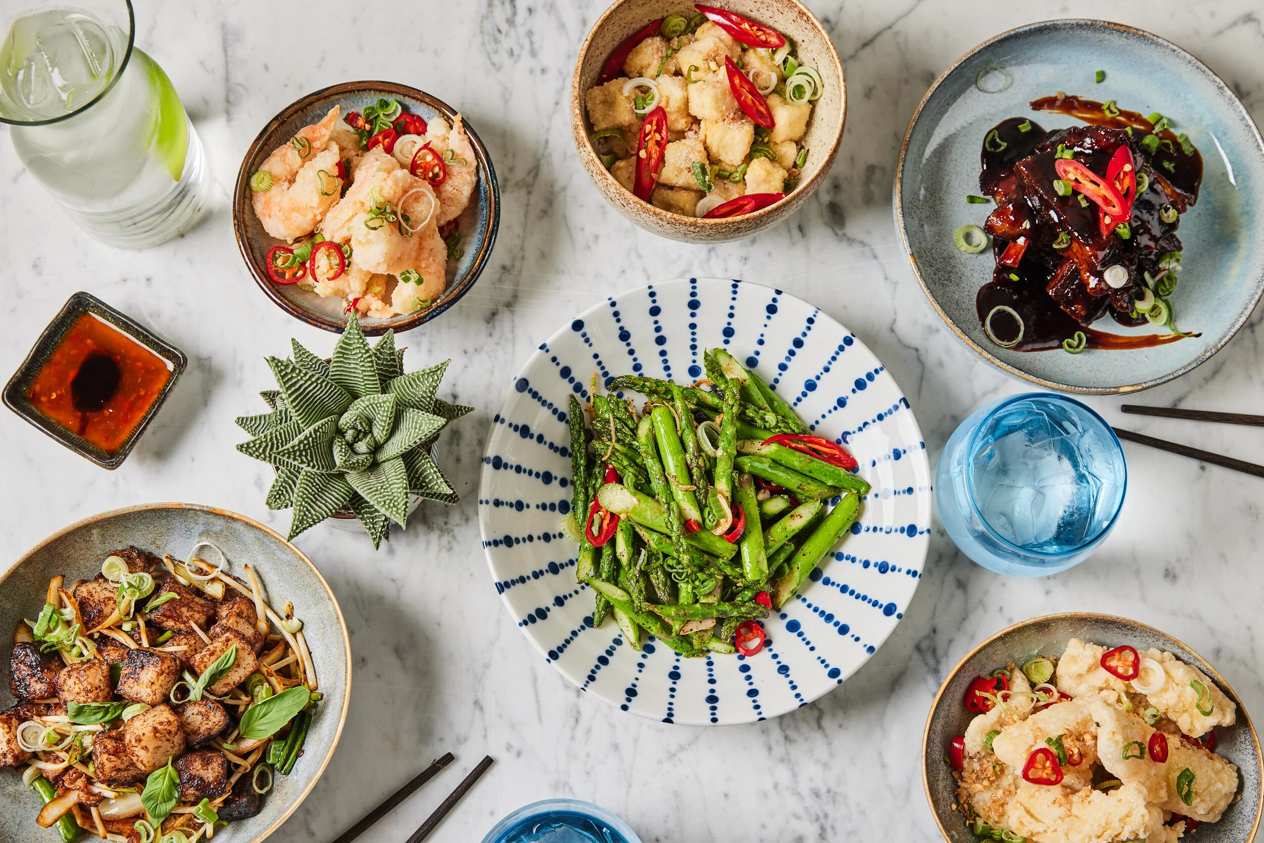 Overhead spread of Asian-inspired dishes at HUŎ, including crispy seafood, glazed ribs, stir-fried asparagus, noodles, dipping sauce, drinks, and chopsticks on a marble table