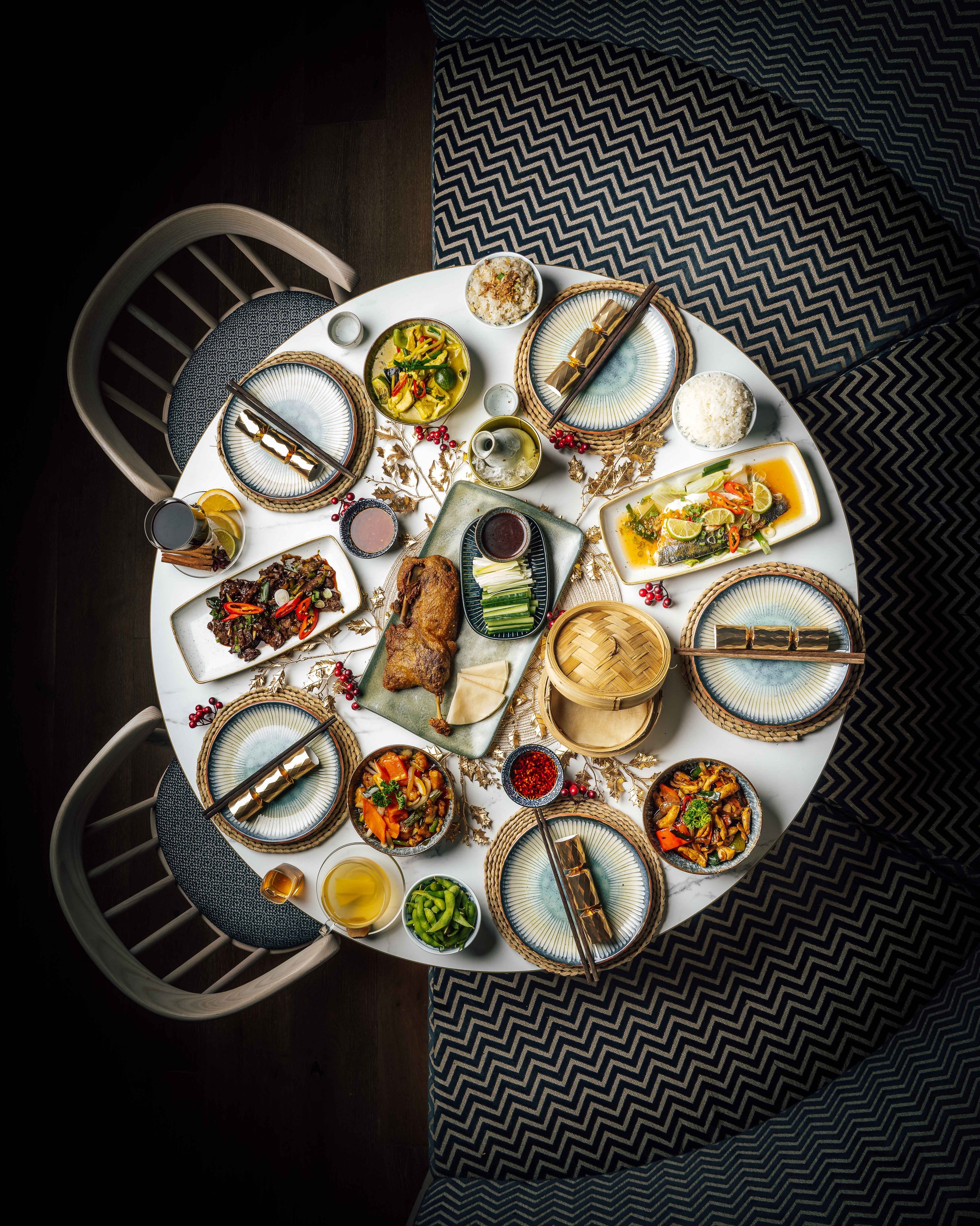 Overhead view of a festive Asian-inspired dinner table at HUŎ with oriental style plates, rice, vegetables, roasted duck, fish, chopsticks, tea, and decorative place settings.