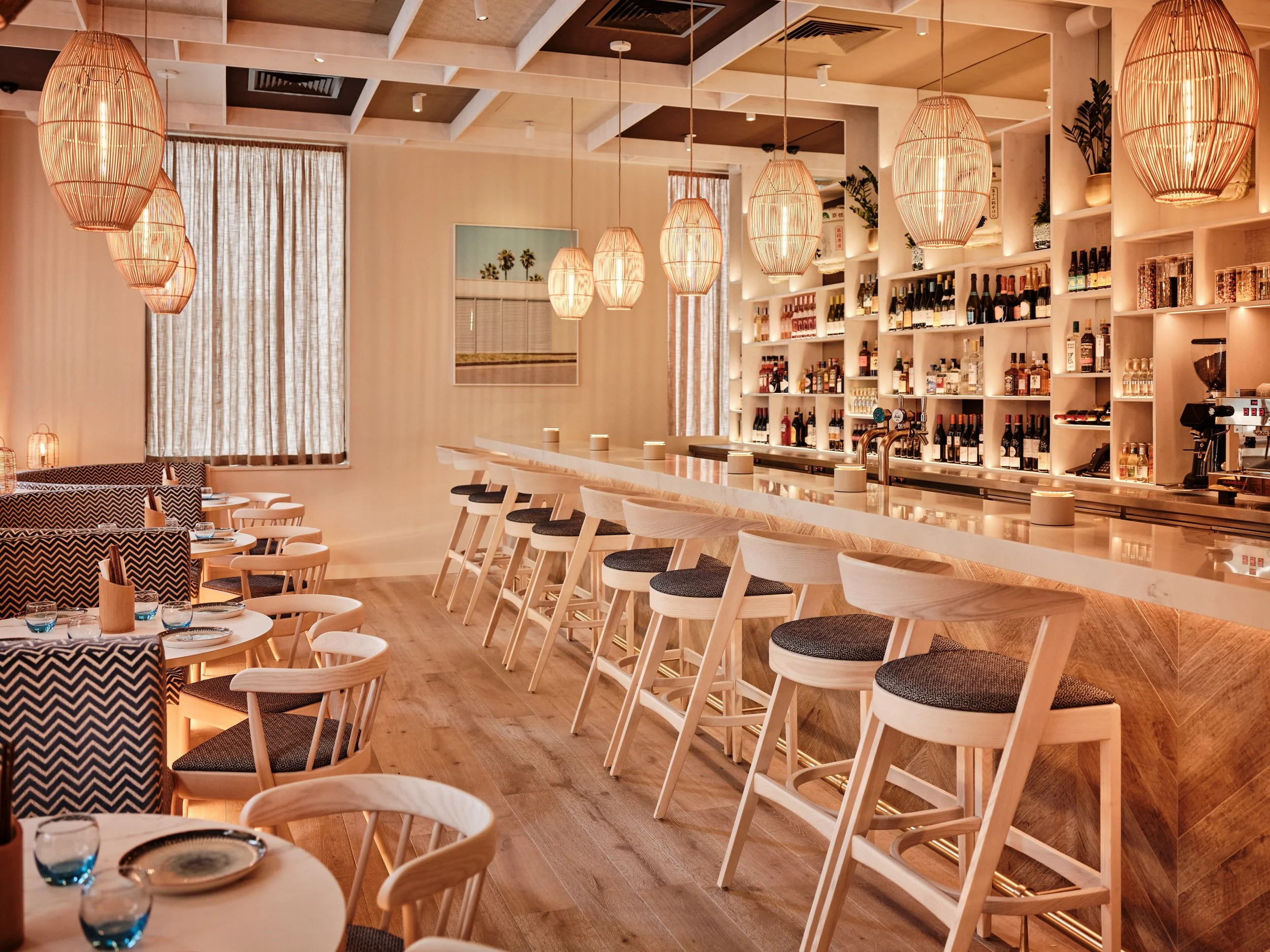 Warmly lit modern restaurant interior with woven pendant lights, bar shelves stocked with wine and spirits, and rows of wooden dining tables and chairs.