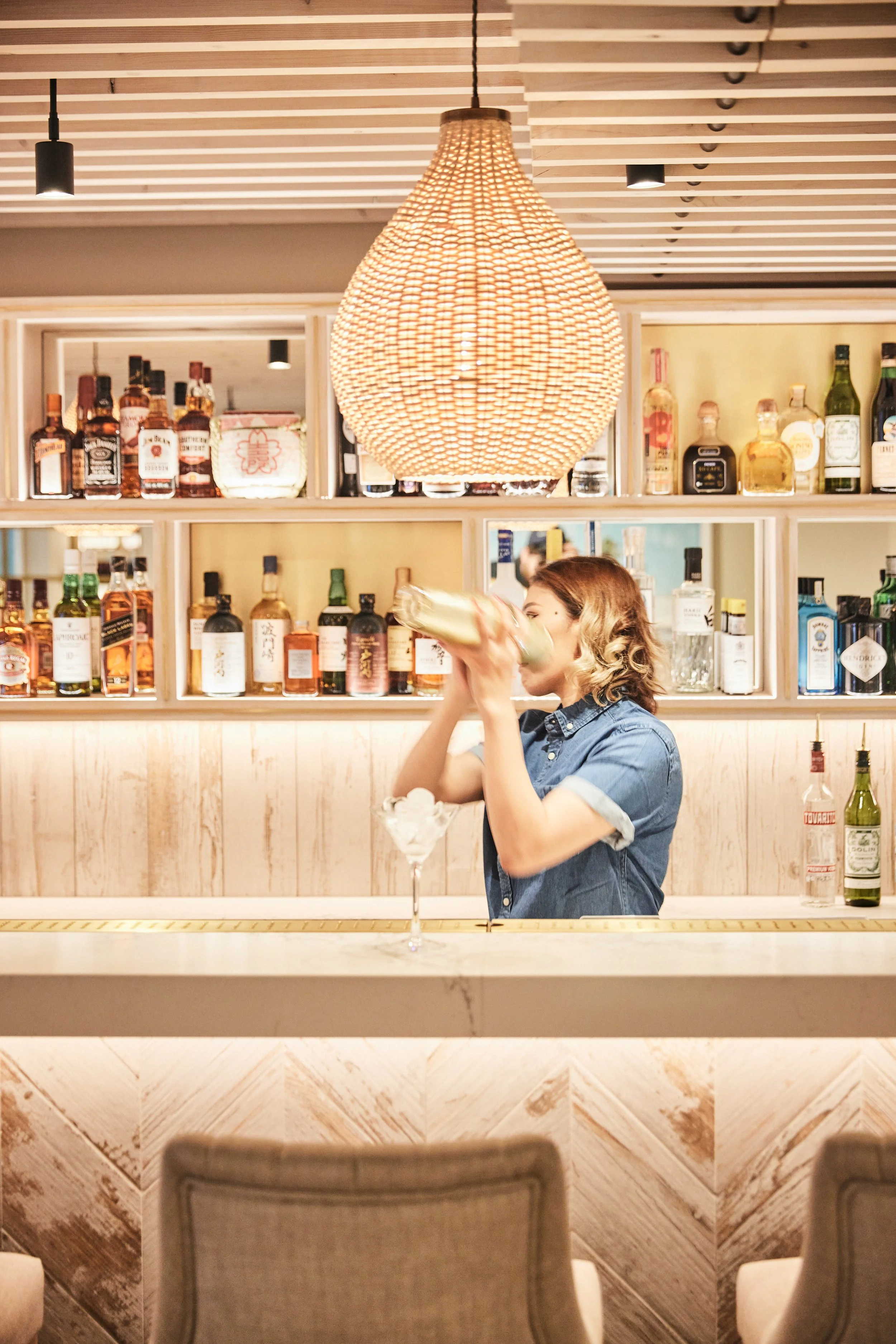 Bartender at HUŎ shaking a cocktail in front of a liquor bottle display at a warmly lit bar