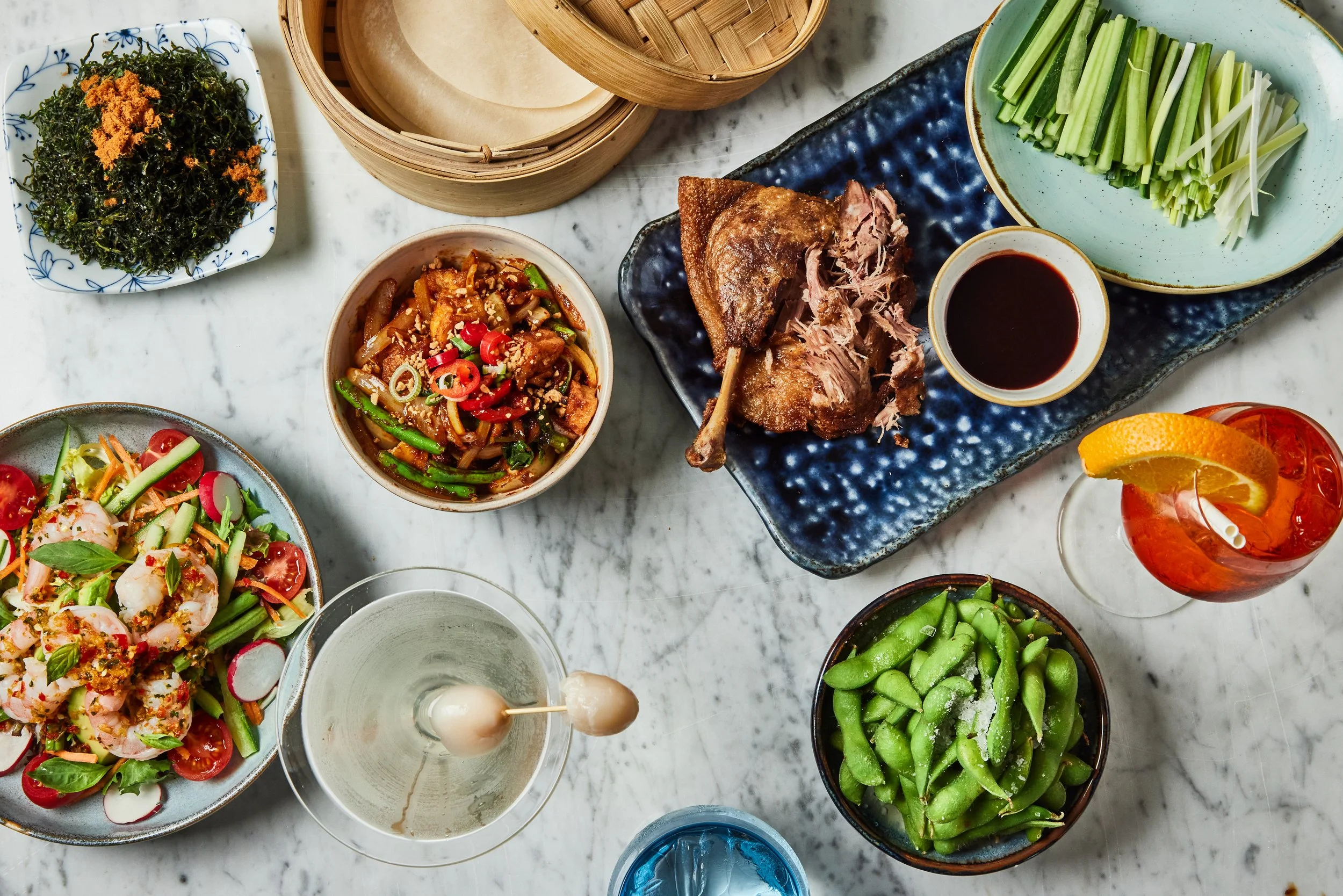 Overhead spread of Asian dishes at HUŎ with roasted duck, rice paper, vegetables, salads, edamame, dipping sauce, and colorful cocktails
