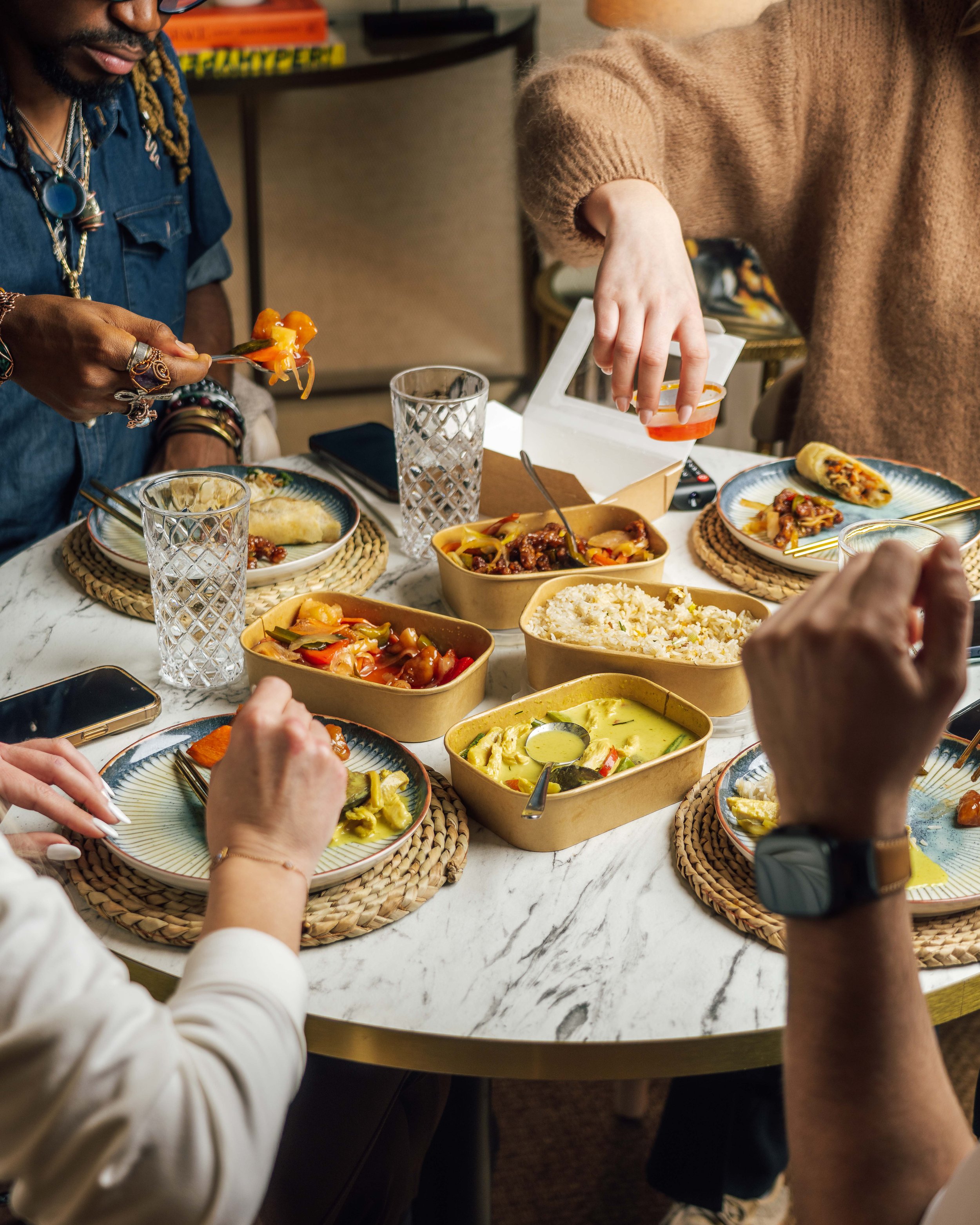 Friends sharing a colorful Asian meal around a dining table with rice, noodles, vegetables, and drinks