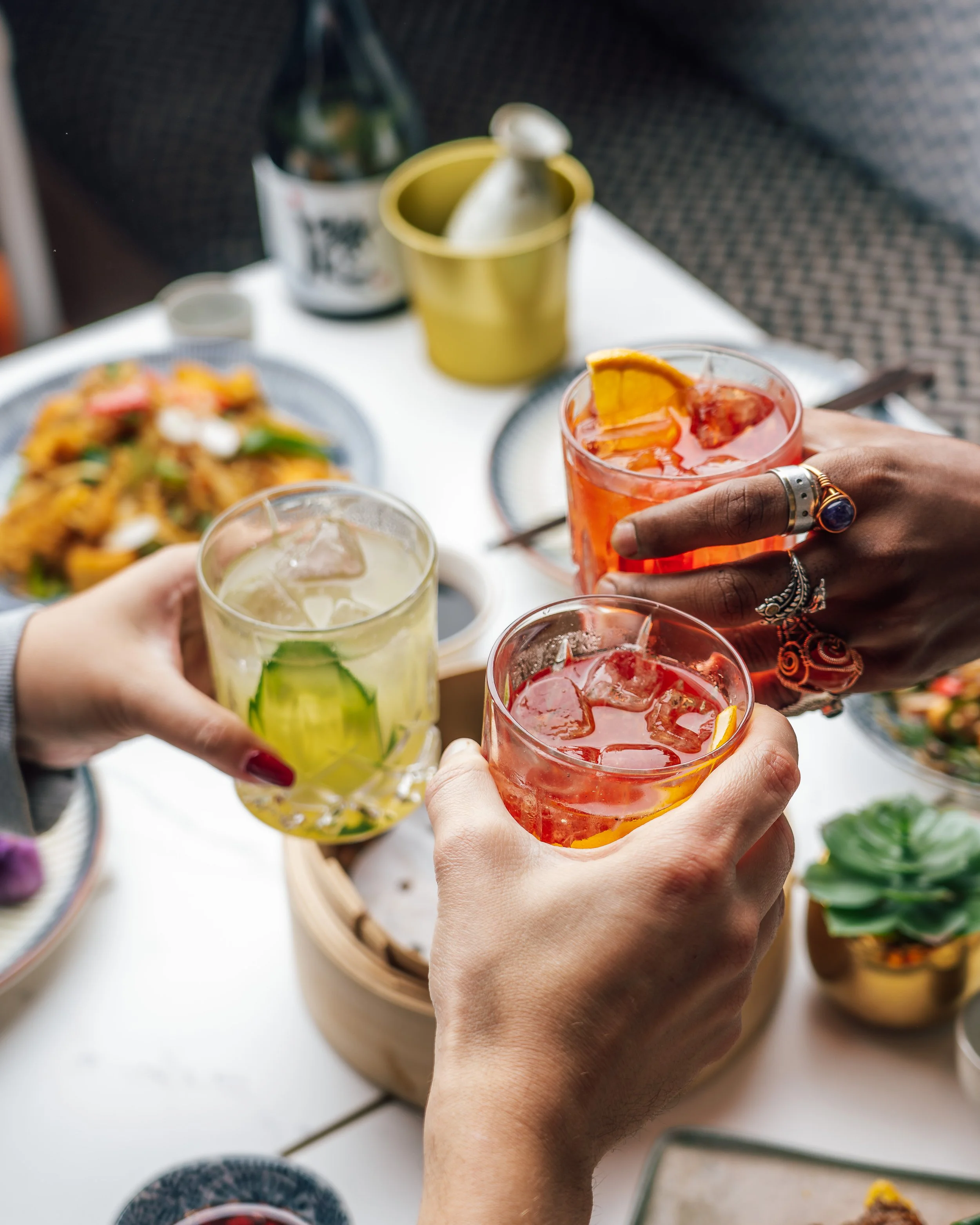 Friends raising colorful cocktails at an outdoor dining table with food at HUŎ