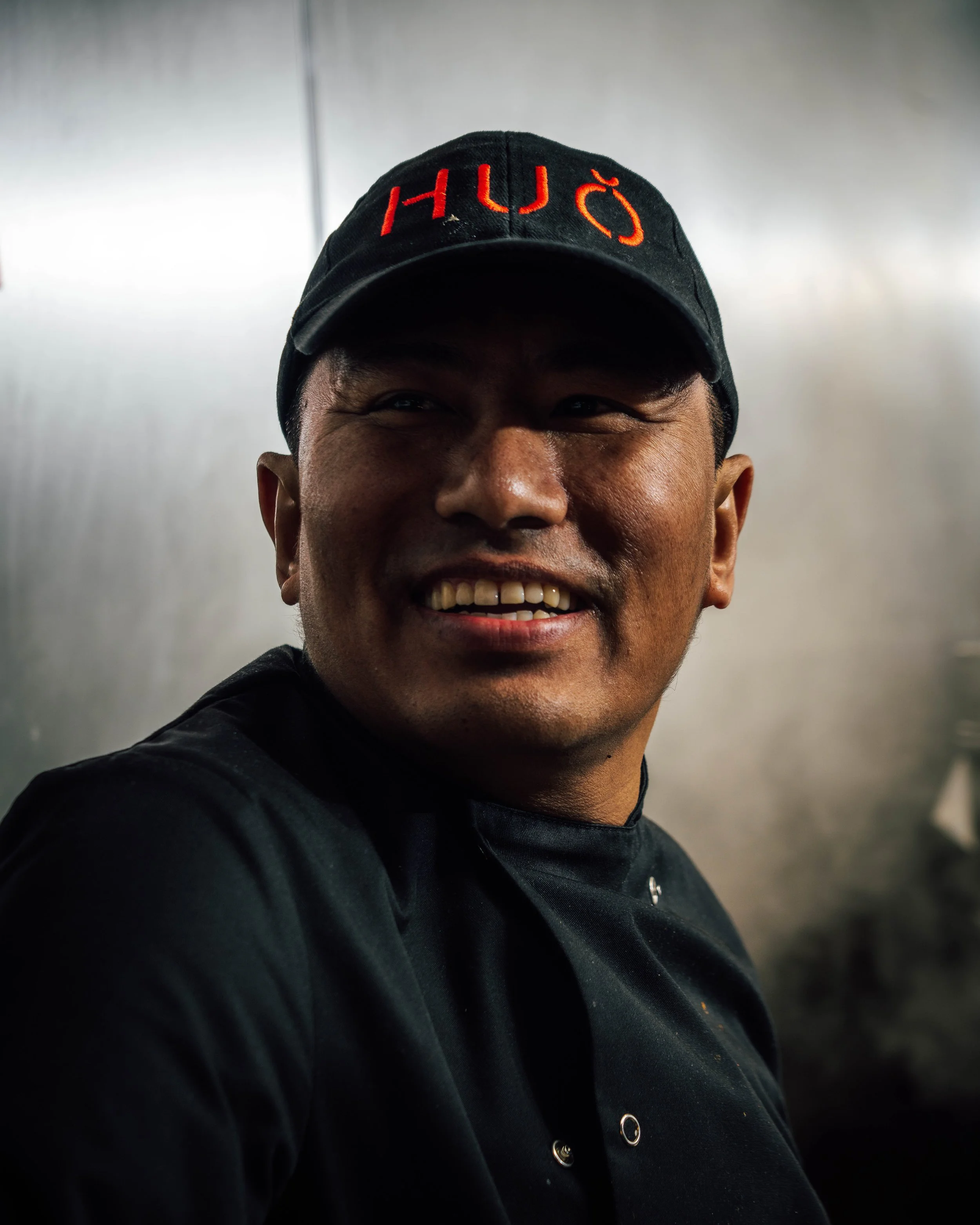 Head Chef Yam at HUŎ smiling man wearing a black cap with red lettering and a black shirt