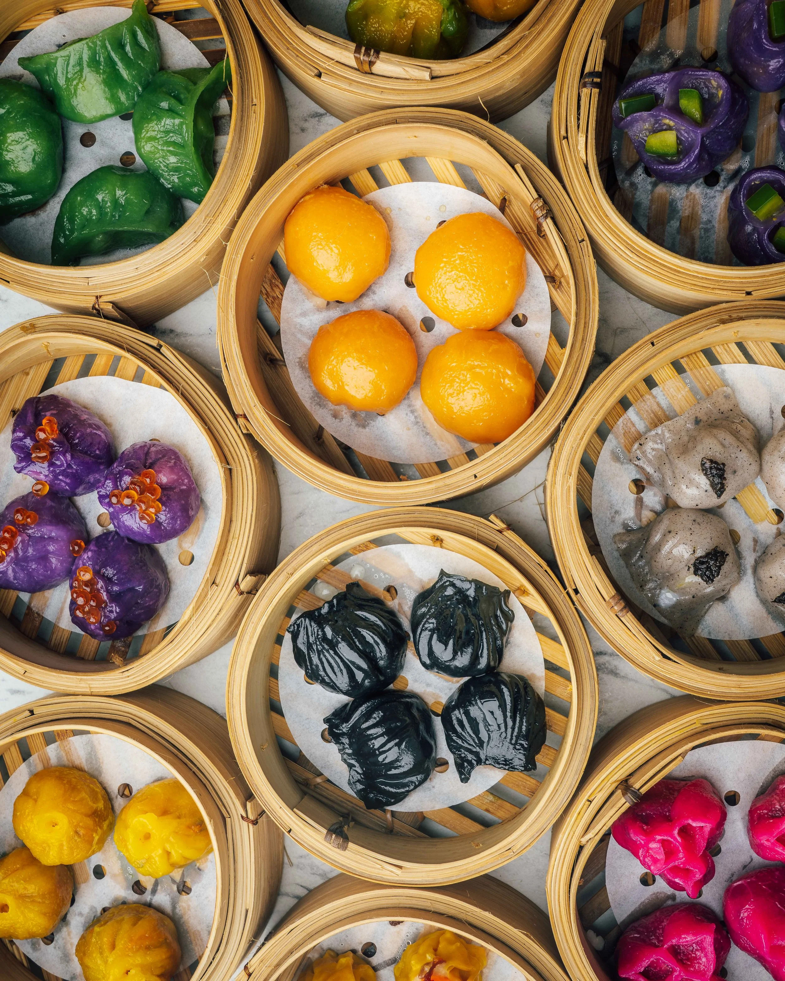 Overhead view of HUŎ's colorful steamed dumplings in bamboo baskets, including orange, green, purple, black, yellow, and pink varieties.