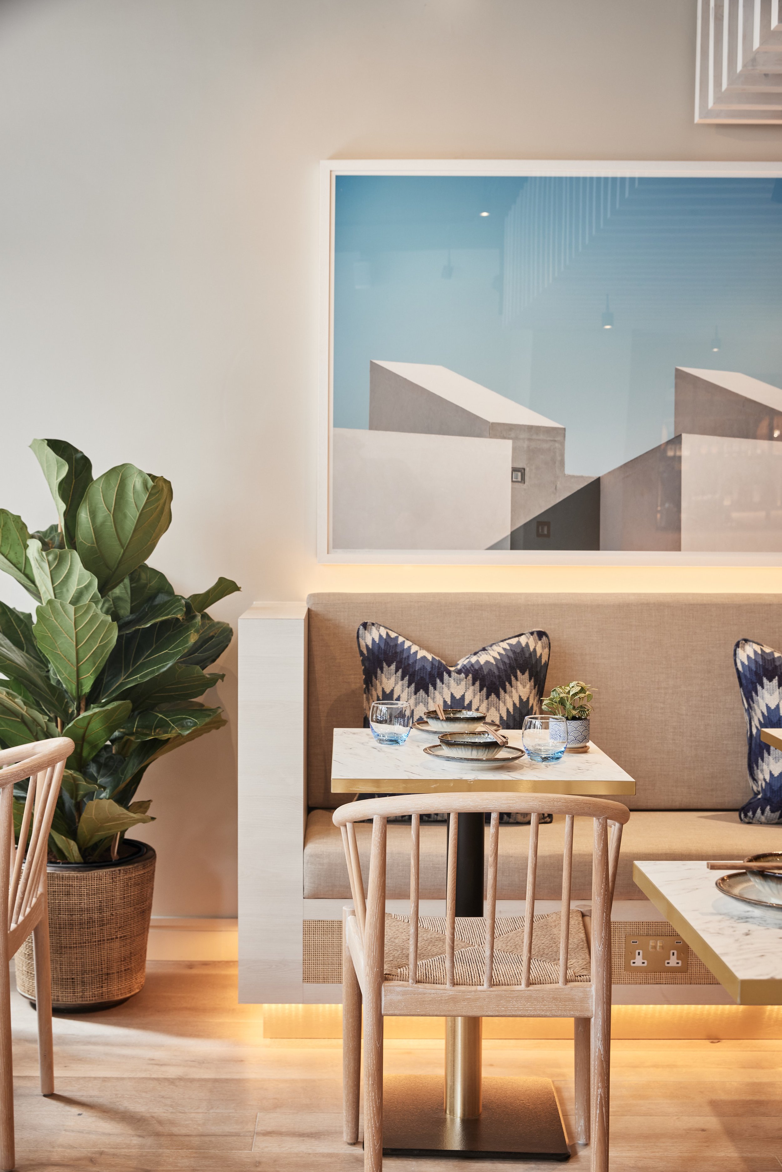 Stylish restaurant dining area at HUŎ Chelsea with a large potted fiddle-leaf fig, upholstered banquette, patterned pillows, set marble tables, and framed abstract artwork.