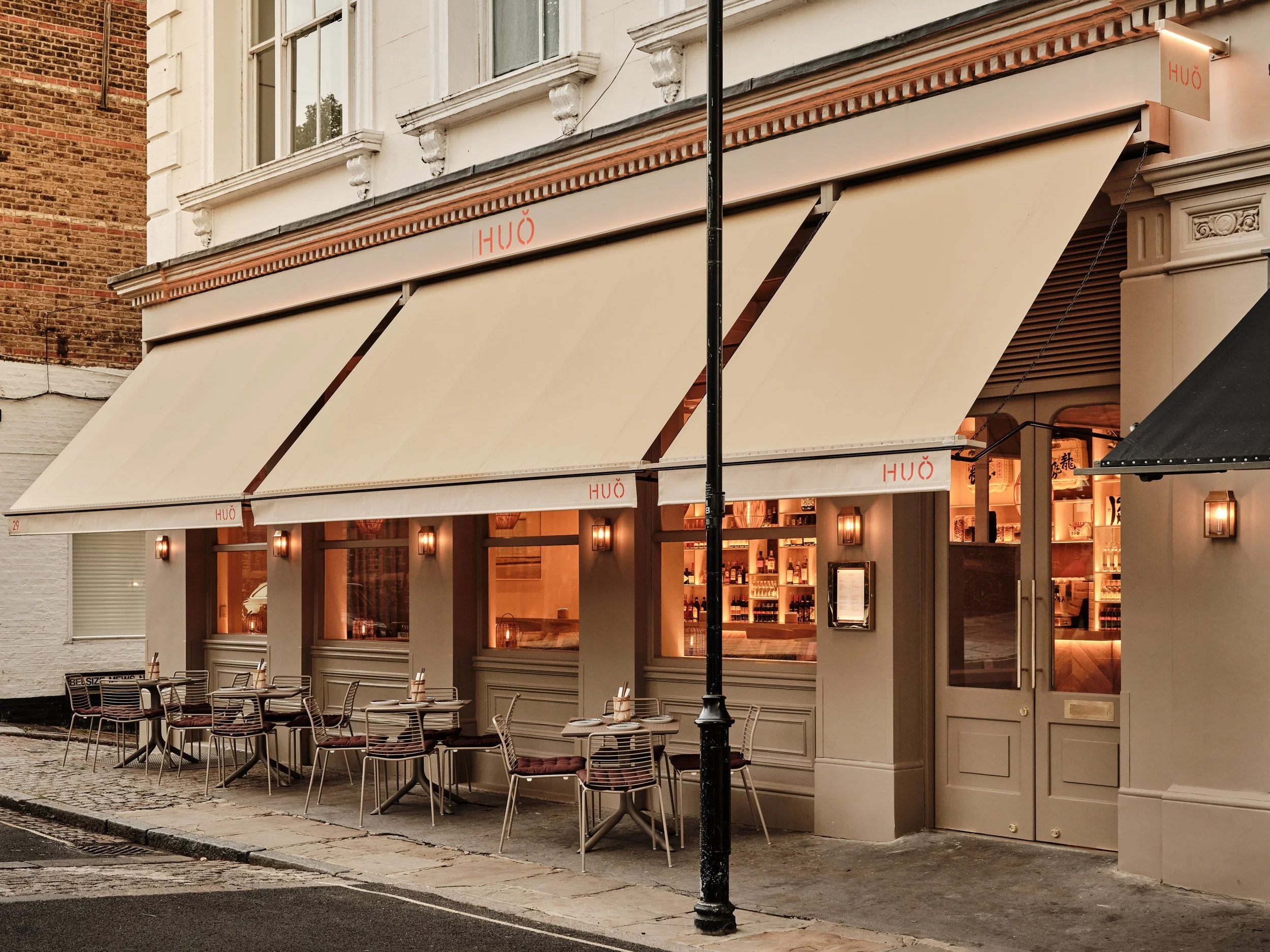 HUŎ restaurant Belsize Village exterior with cream awnings, outdoor tables, and lantern-lit dining room on a cobblestone street
