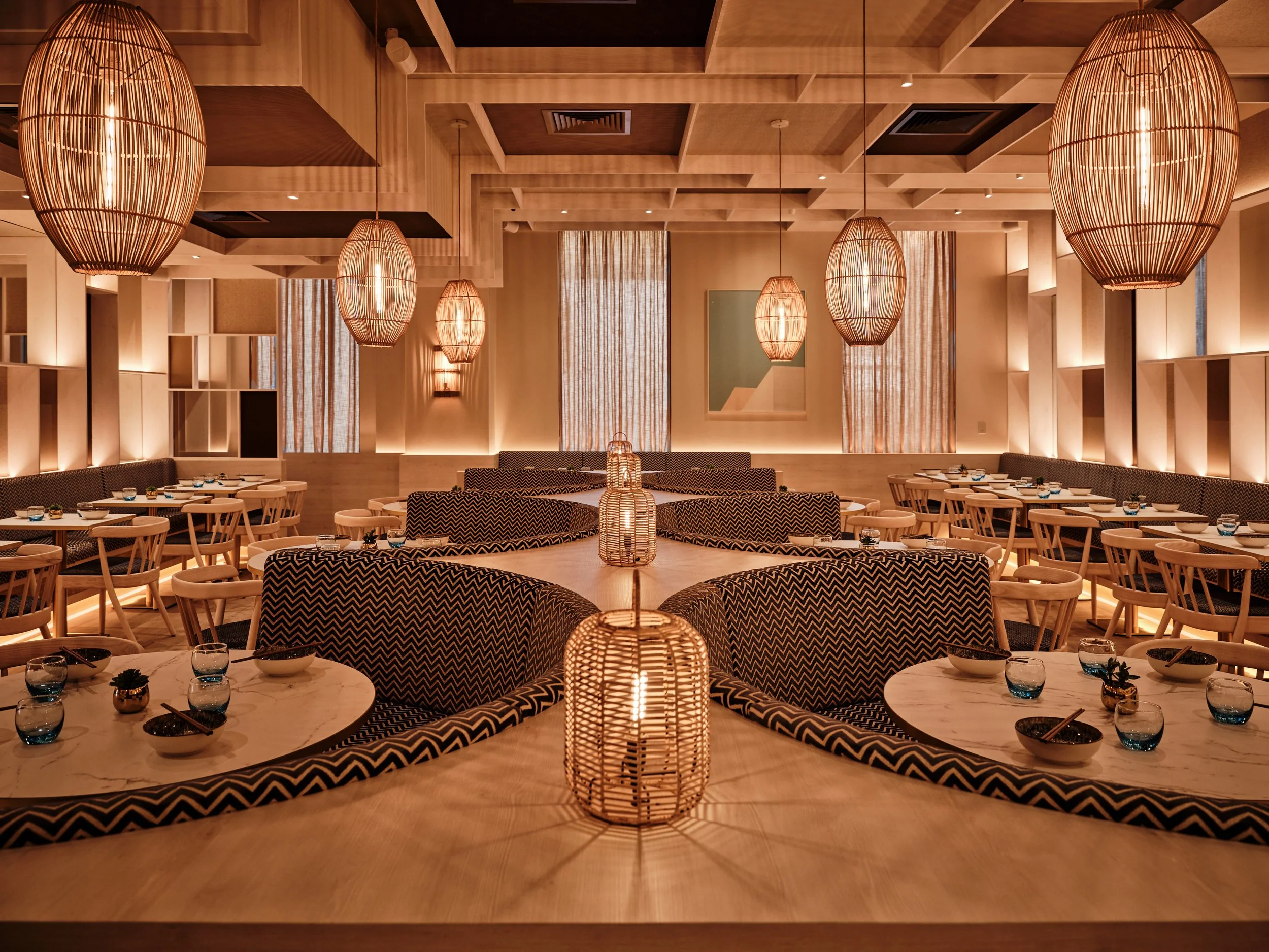 Warmly lit modern restaurant interior of HUŎ Belsize Village with woven pendant lights, patterned banquette seating, and neatly set dining tables.