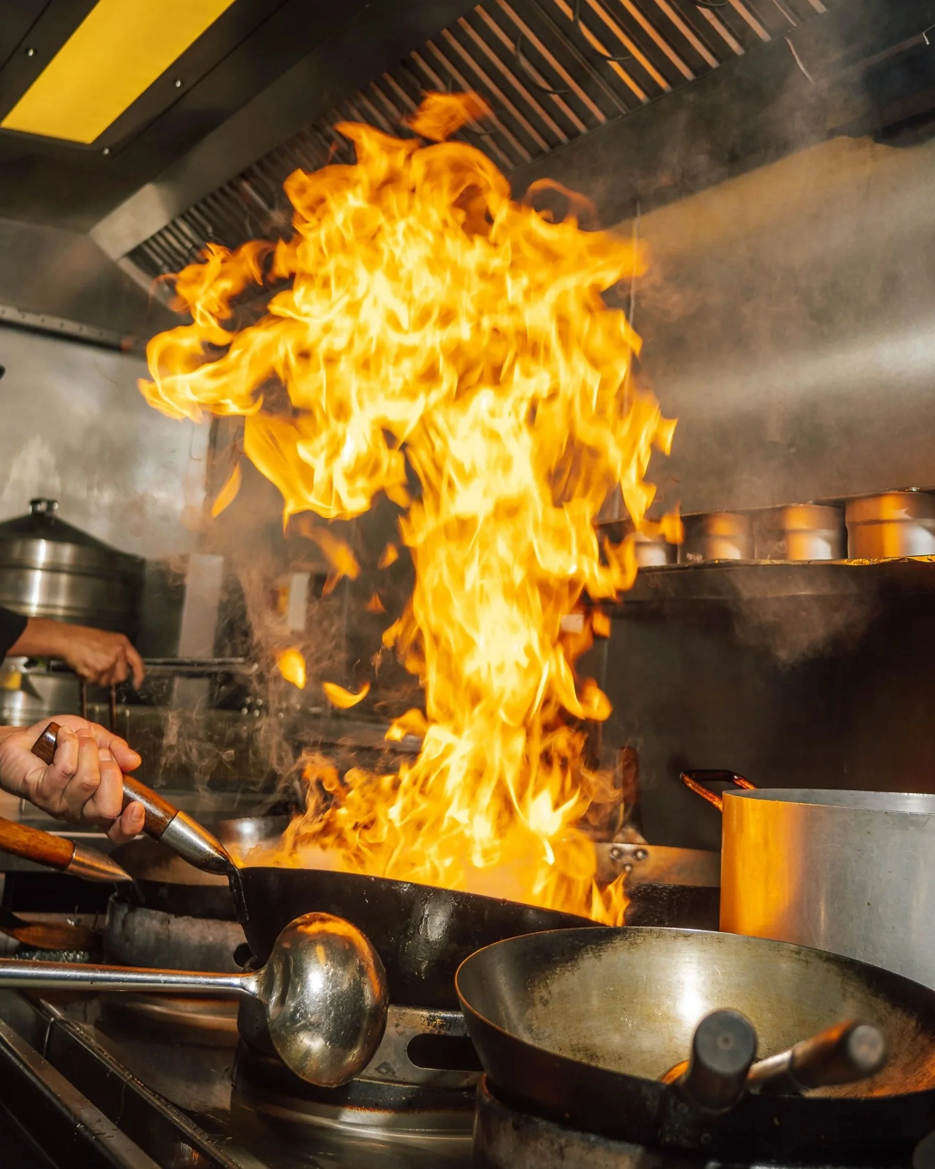 Chef at HUŎ cooking in a commercial kitchen with a large wok engulfed in flames