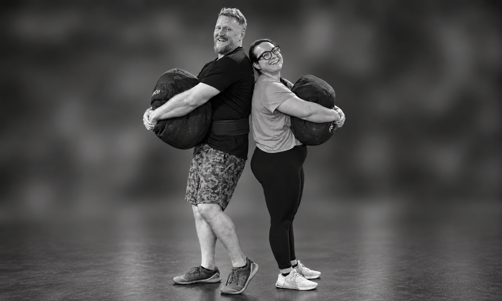 Two people, a man and a woman, standing back-to-back, each holding a large weighted sandbag in front of them, smiling and laughing. The man has short, light-colored hair and a beard, wearing a black shirt and camouflage shorts. The woman has dark hair tied back, wearing glasses, a light T-shirt, and black leggings.