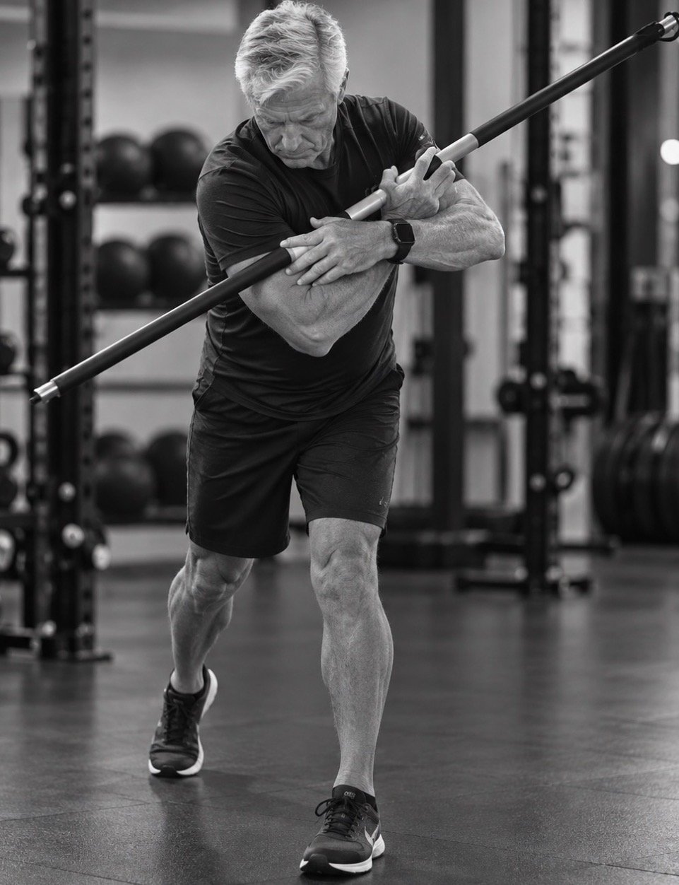 An older man working out in a gym, holding a bar across his shoulders, preparing for a squat or similar exercise.