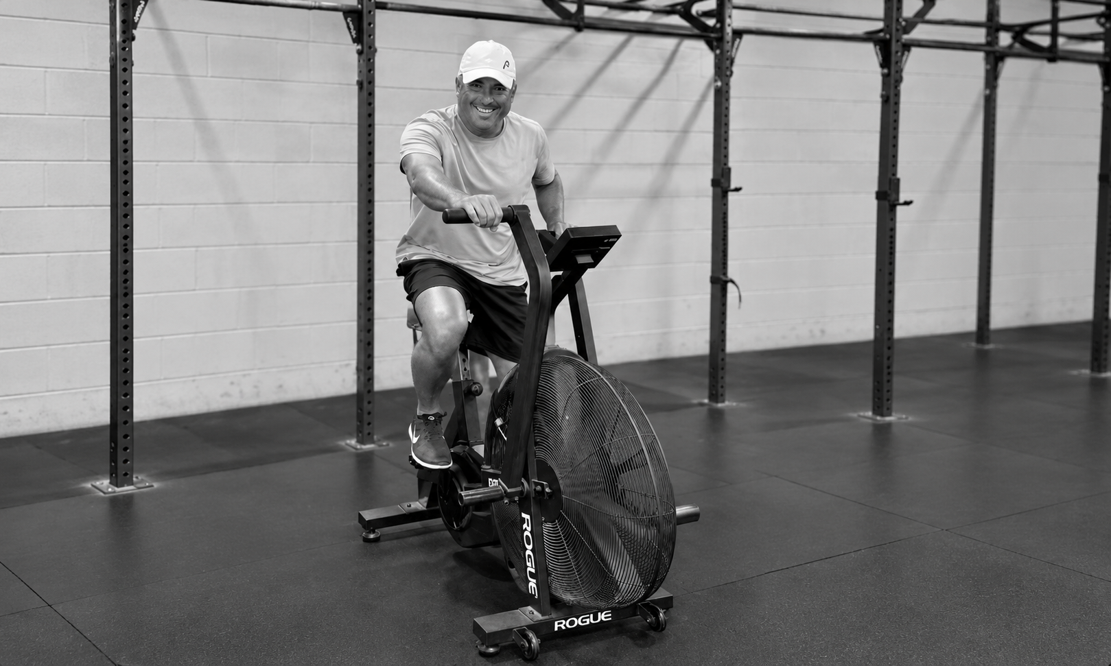 A man smiling while riding a stationary exercise bike with a large fan at a gym.
