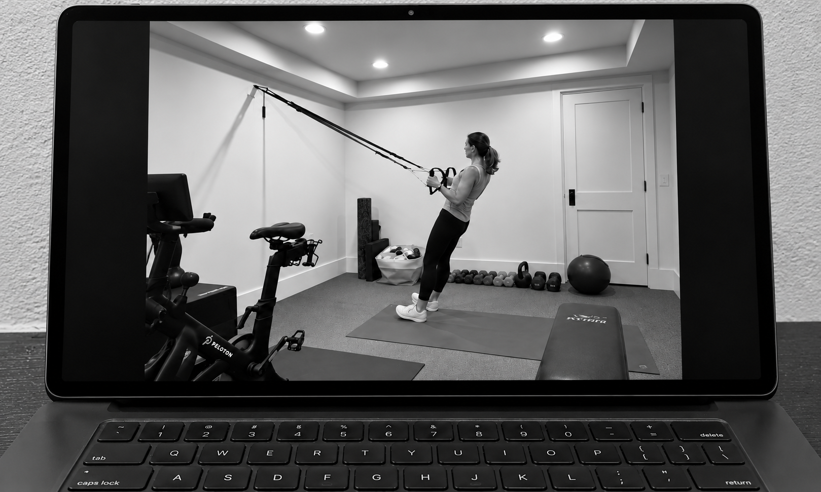 A woman is exercising with resistance bands attached to the wall in a home gym, with various workout equipment such as a stationary bike, weights, and a stability ball around her.