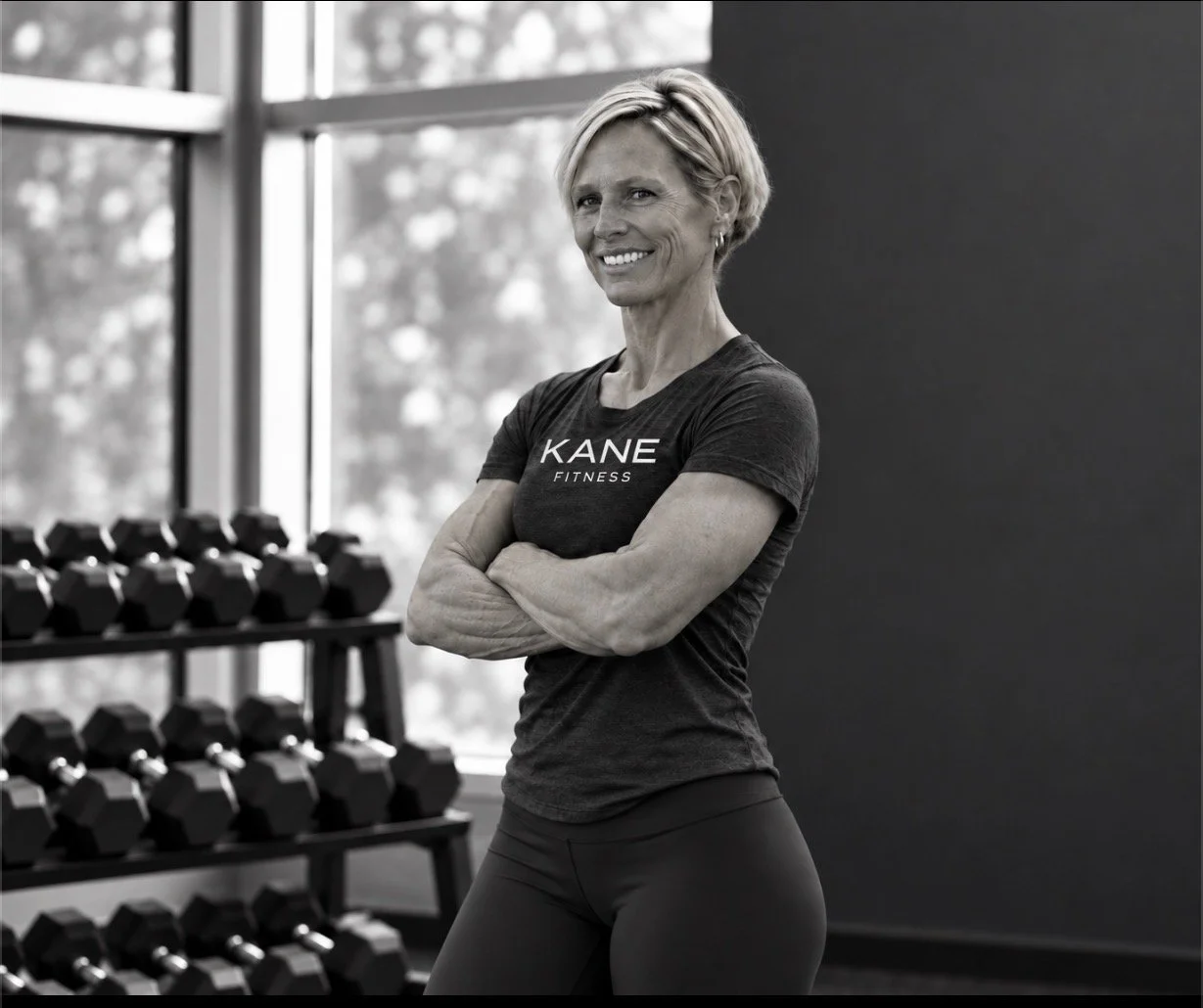 A smiling mature woman with short blonde hair, wearing a dark t-shirt with 'KANE FITNESS' written on it, posing with crossed arms in a gym near a rack of black dumbbells and large windows.