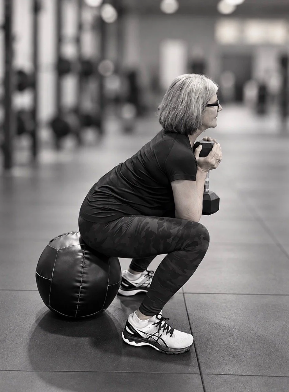 A woman with gray hair, glasses, and sportswear, sitting on a medicine ball while holding a dumbbell close to her chest in a gym.