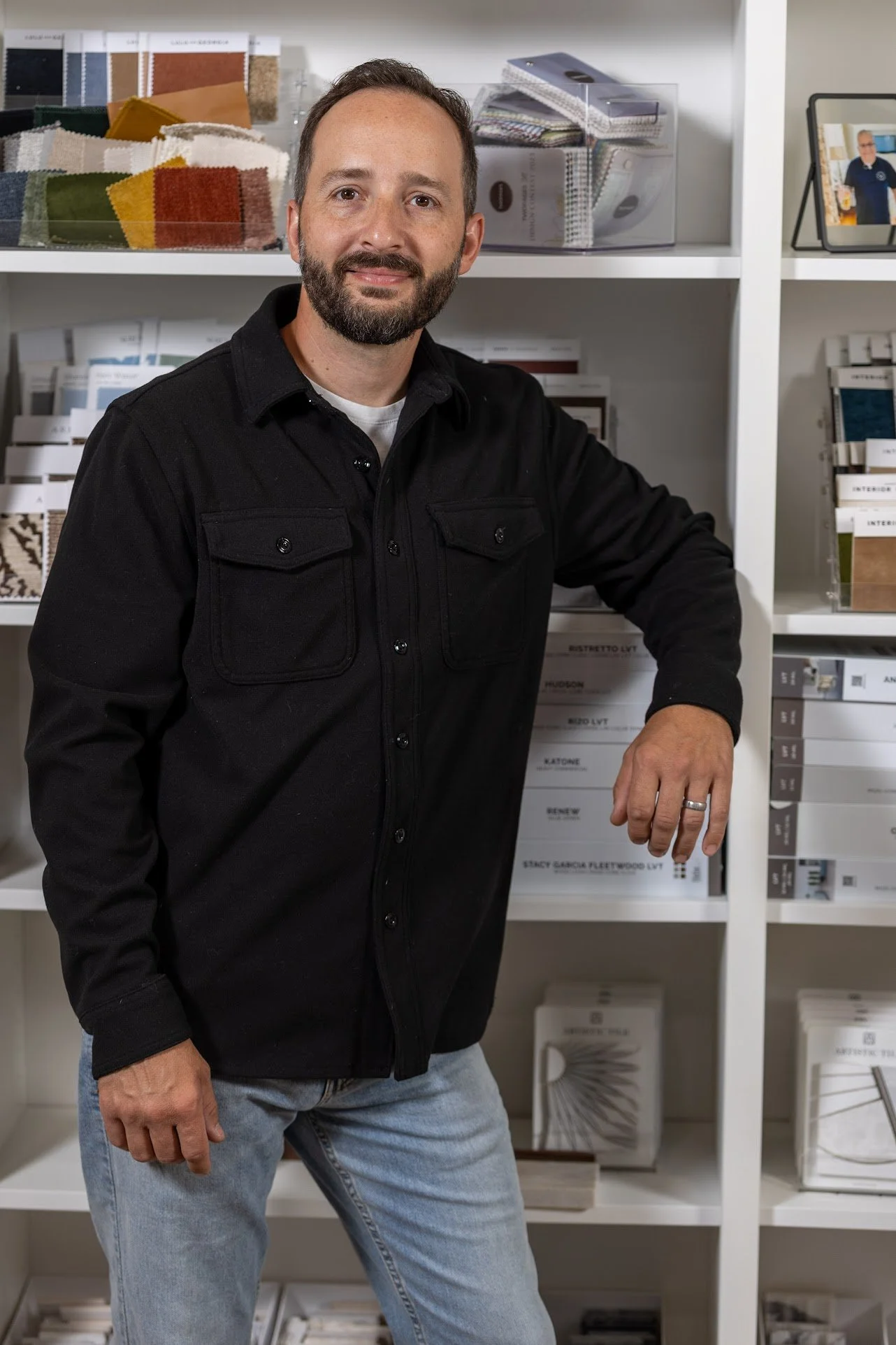 Man with beard wearing black jacket and blue jeans standing in an interior design studio, with shelves of fabric samples, books, and magazine stands in the background.
