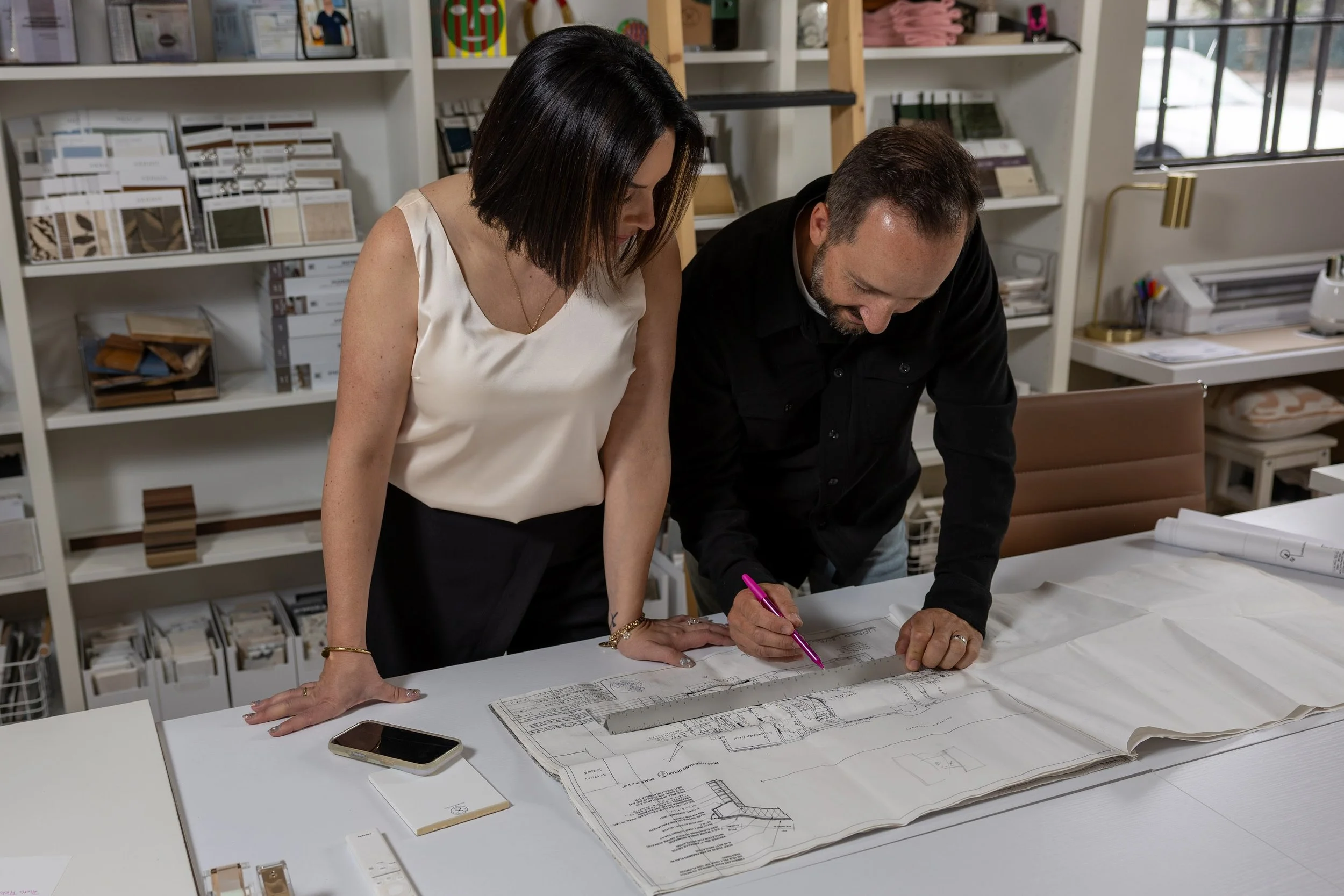 Two people examining architectural blueprints at a table in an office or design studio. Shelves in the background hold various design samples and books.
