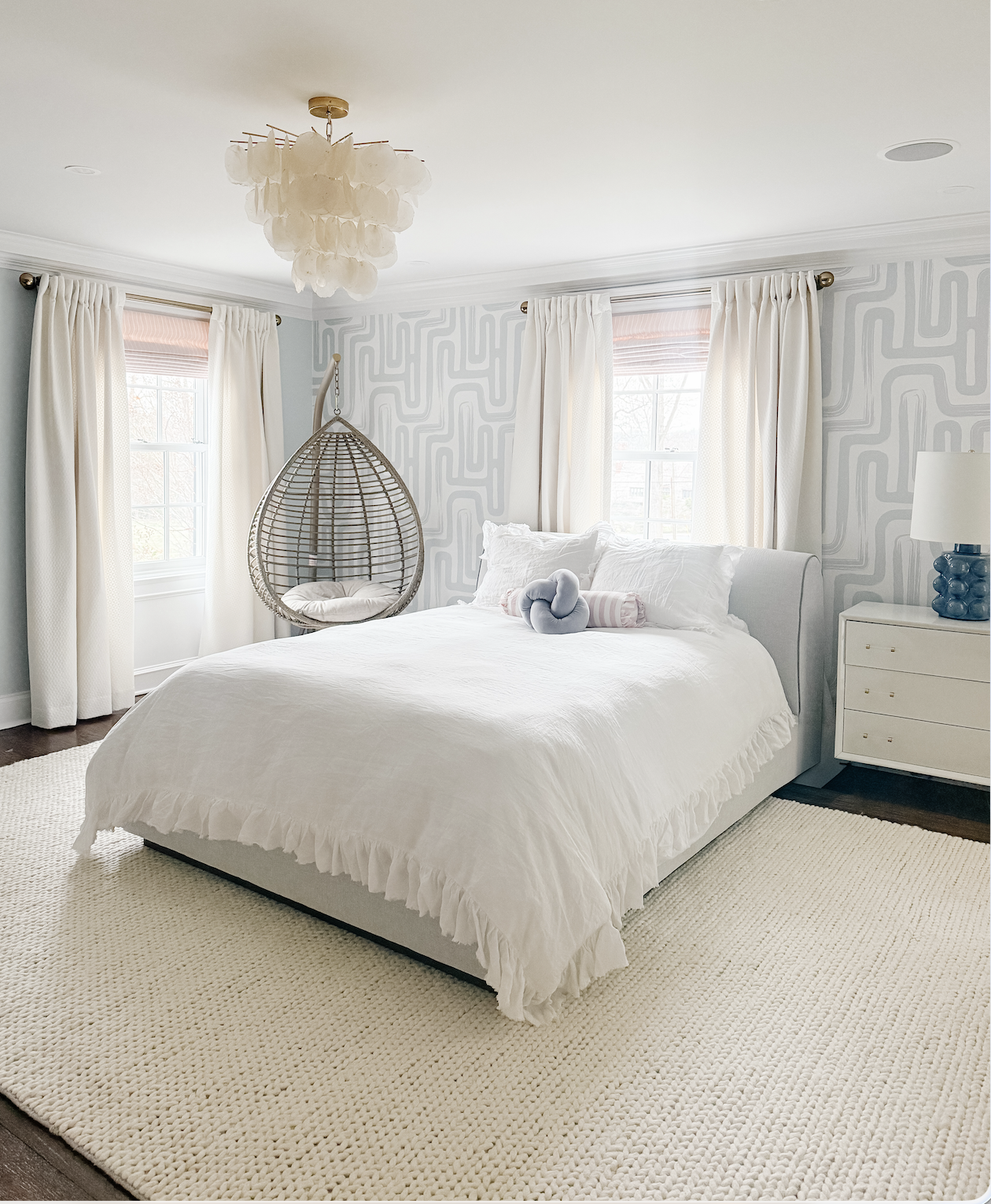 A bright and airy bedroom with white bedding, a hanging wicker chair, and light-colored curtains. There's a white nightstand with a blue lamp and a plush knot pillow on the bed. The room has a patterned accent wall, a chandelier, and a textured area rug.
