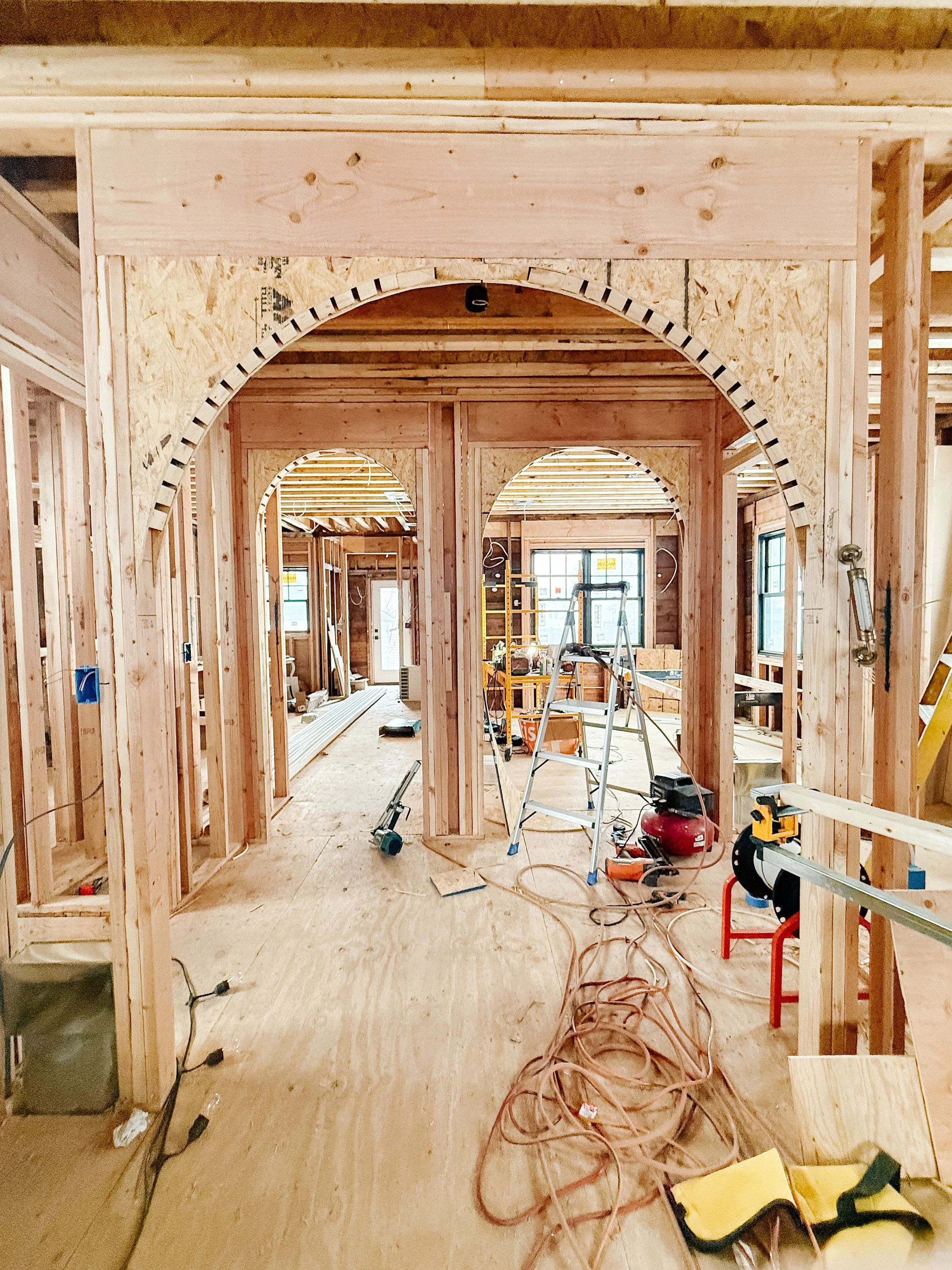 Interior view of a house under construction, showing wooden framing, arches, and construction tools and equipment scattered throughout the space.