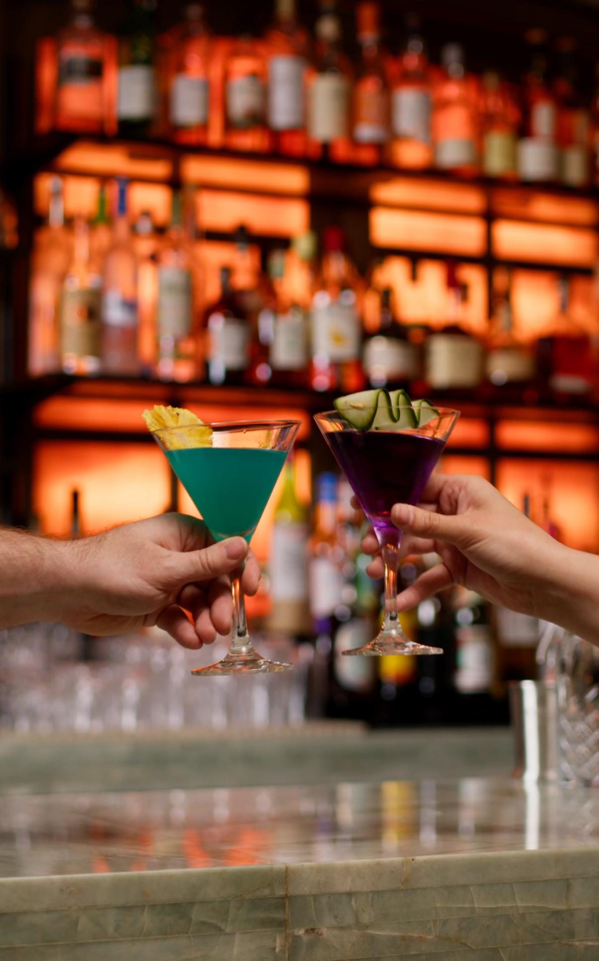 Two people holding colorful cocktails in martini glasses at a bar, with a background of liquor bottles on illuminated shelves.