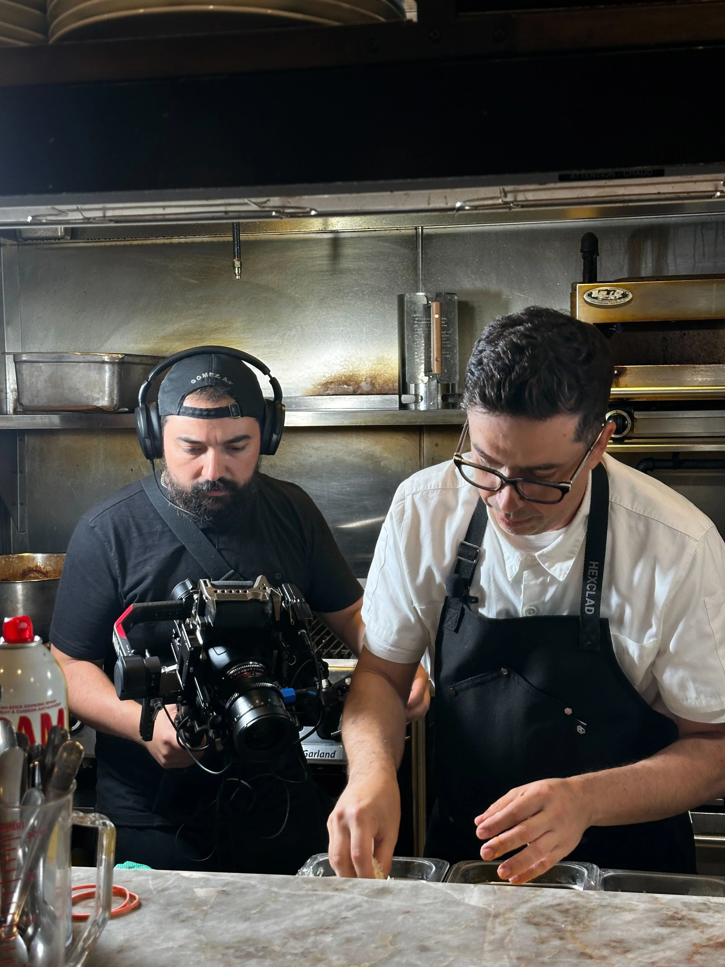 Two men working in a kitchen, one wearing a black shirt, a backward baseball cap, and headphones, holding camera equipment, and the other wearing glasses, a white shirt, and a black apron, preparing food on a countertop.