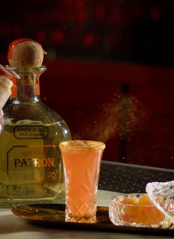 A cocktail glass being garnished with a cinnamon stick, with a bottle of Patrón Reposado tequila and a bowl of oranges in the background.